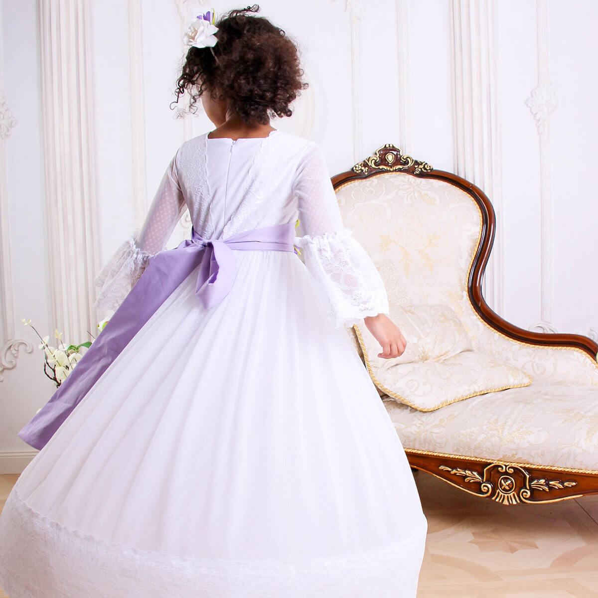 Child in a white dress with a purple sash standing in a decorated room.