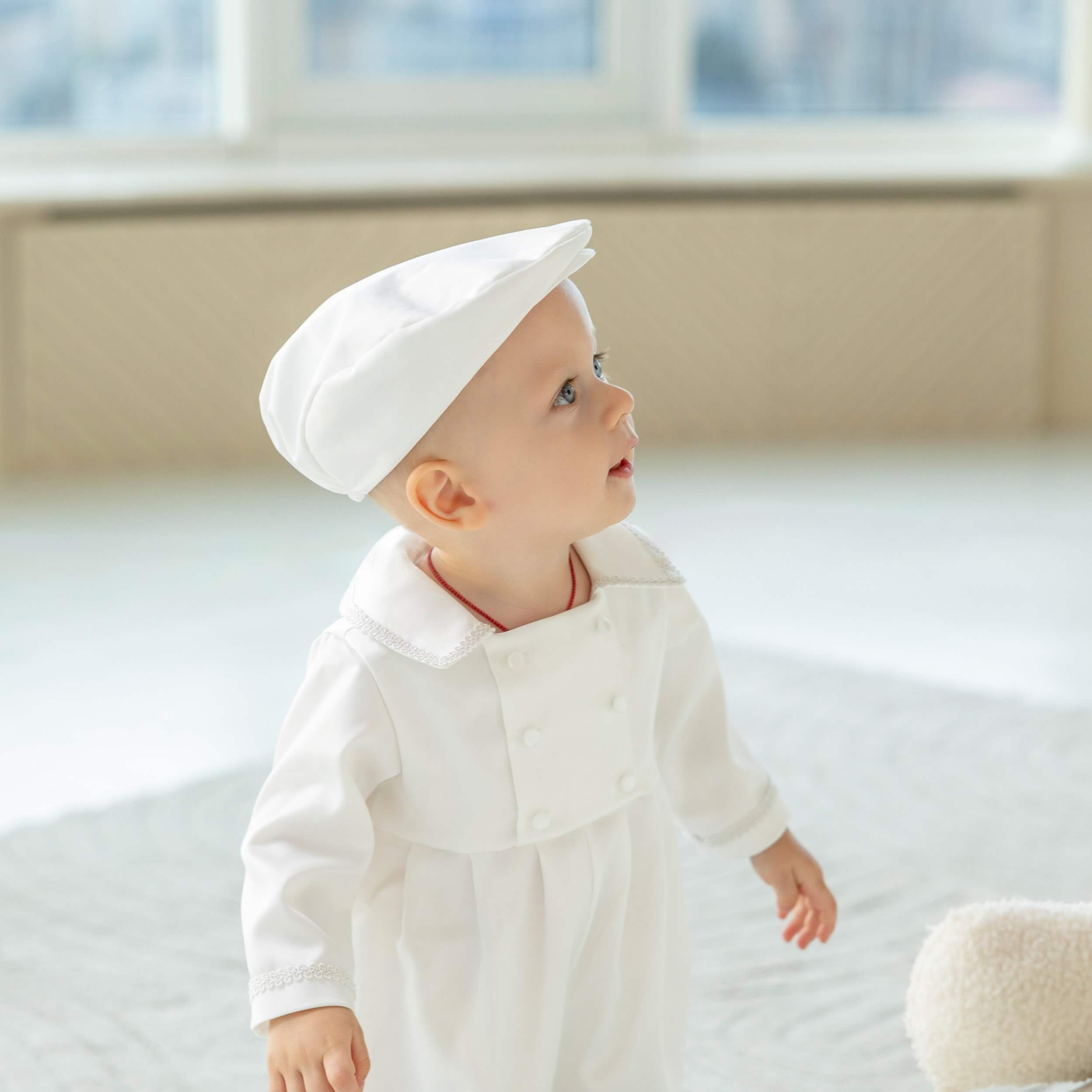 Baby in a Baby Boy Christening Romper and cap standing on a bed with a window in the background