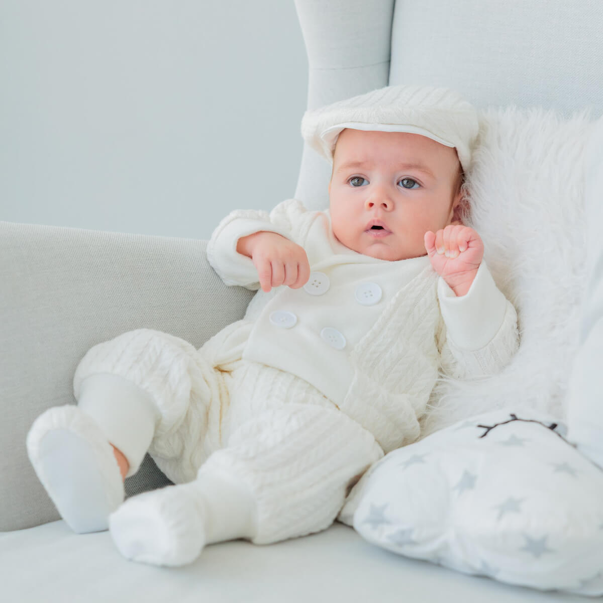 Baby in a Baby Boy Christening Suit sitting on a couch with a soft toy.