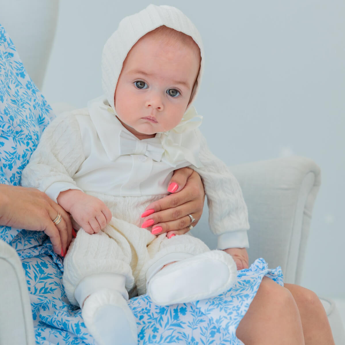 Baby in a Boy Baptism Hat being held by an adult on a floral-patterned chair.