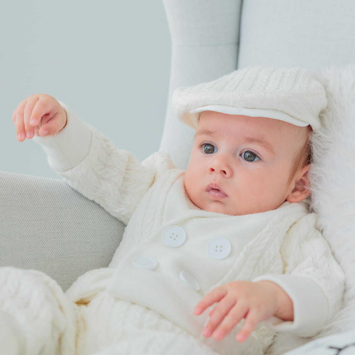 Baby in a white outfit and Boy Christening Hat sitting on a light-colored couch.