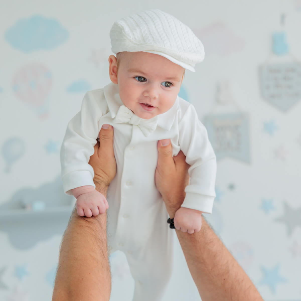 Baby in a white outfit and hBoy Christening Hat held by two hands against a light blue background with cloud patterns.