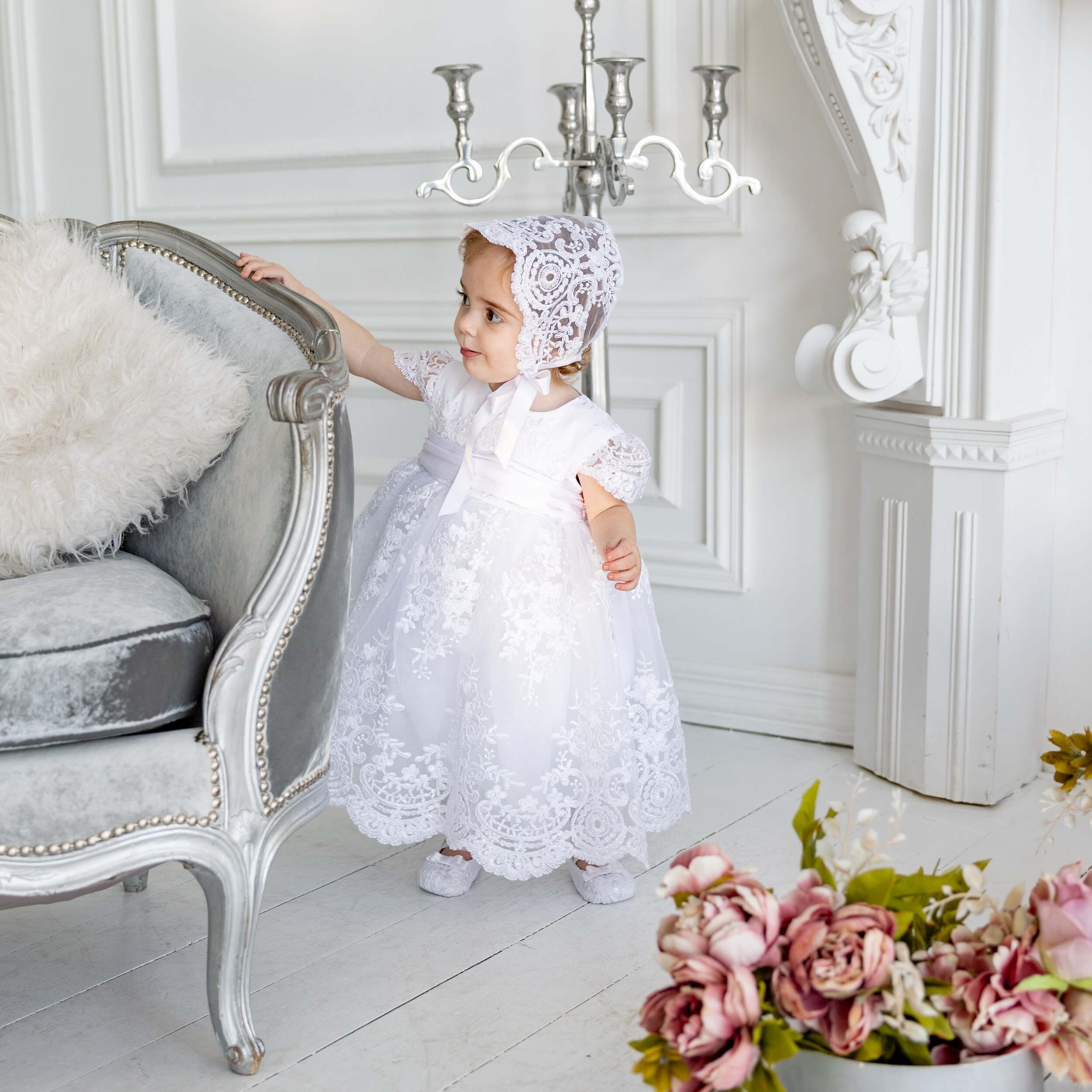 Young girl in a Baptism Dress Set for Girls and bonnet standing in a elegant room with floral decorations.