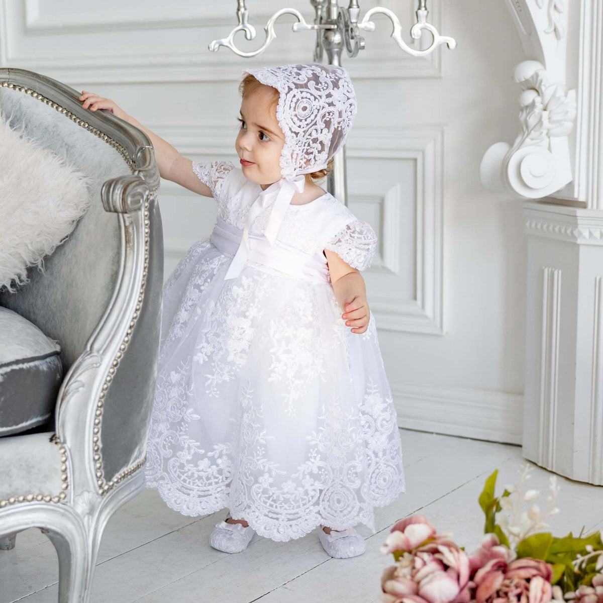 Young girl in a white lace dress and bonnet standing in a elegant room with floral decorations.