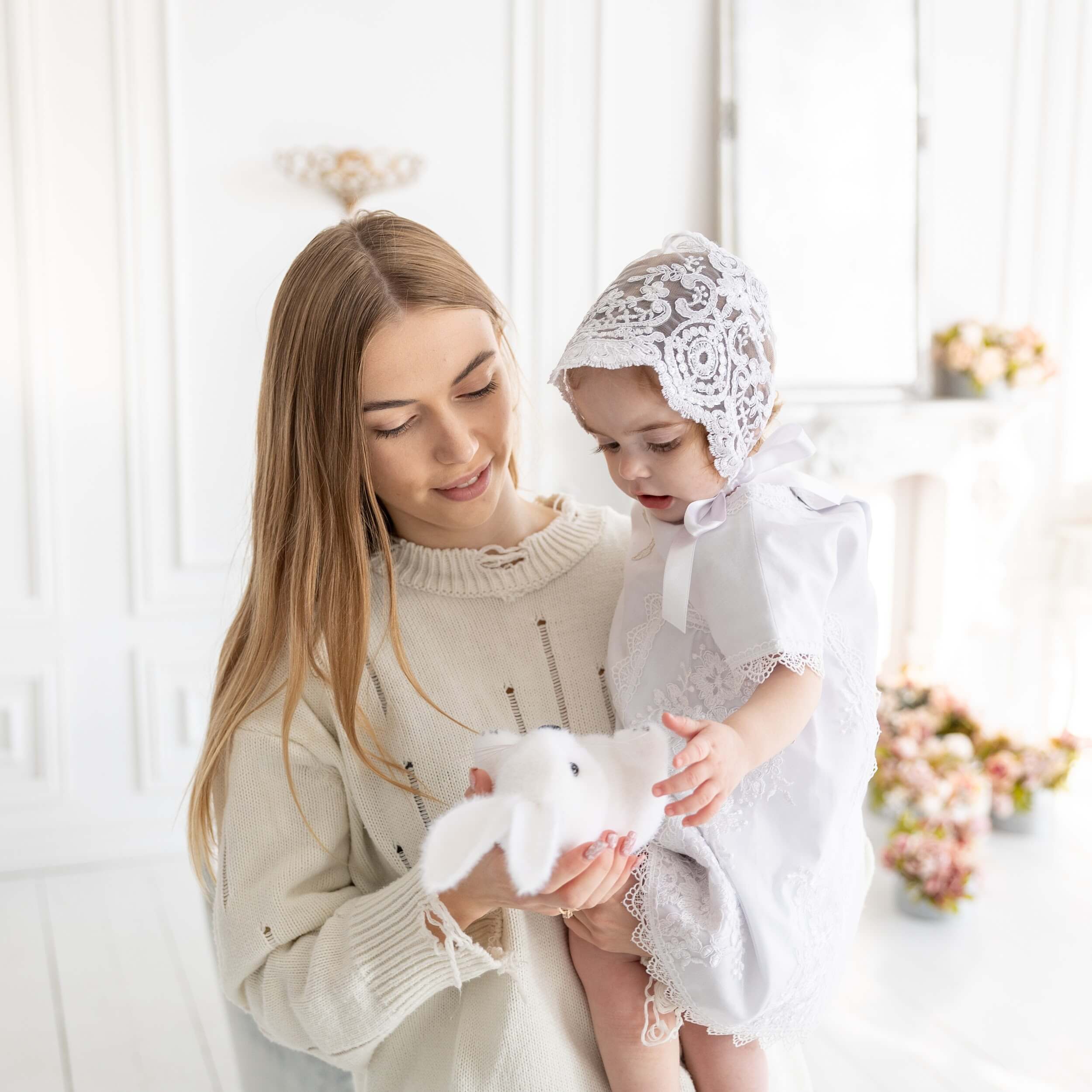 Woman holding a baby in a white lace outfit with a silk christening bonnet in a bright room.