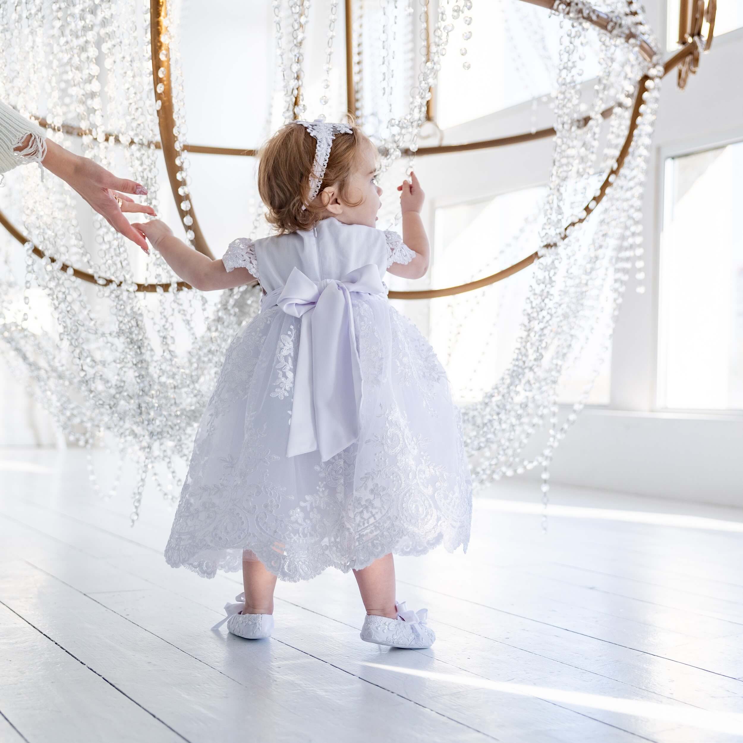 Child in a Baptism Dress for Girls standing on a wooden floor with decorative lights in the background