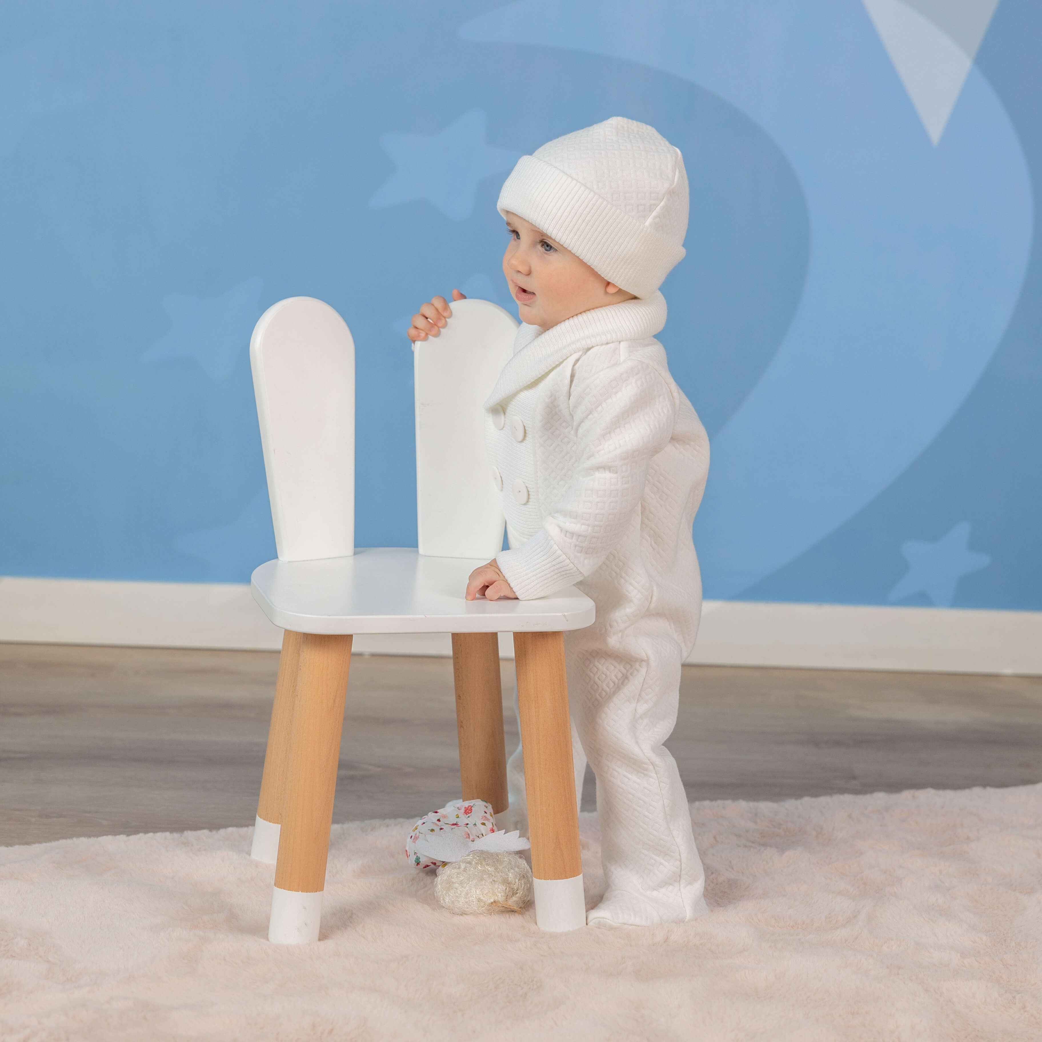 Baptism Outfit for Boys standing next to a white chair with wooden legs against a blue wall.