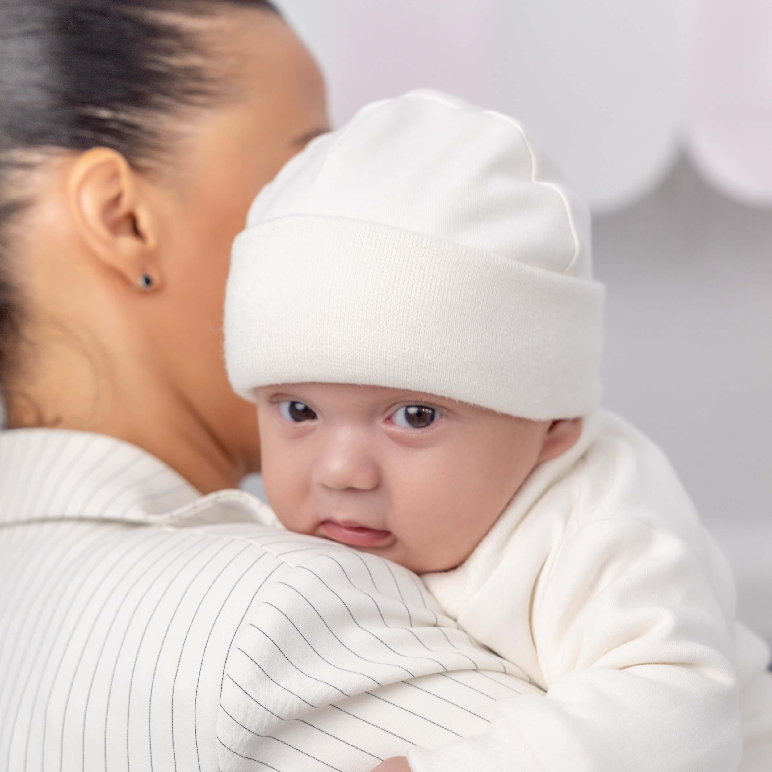 Baby wearing a Boy Christening Hat and wrapped in a white blanket, looking at the camera.