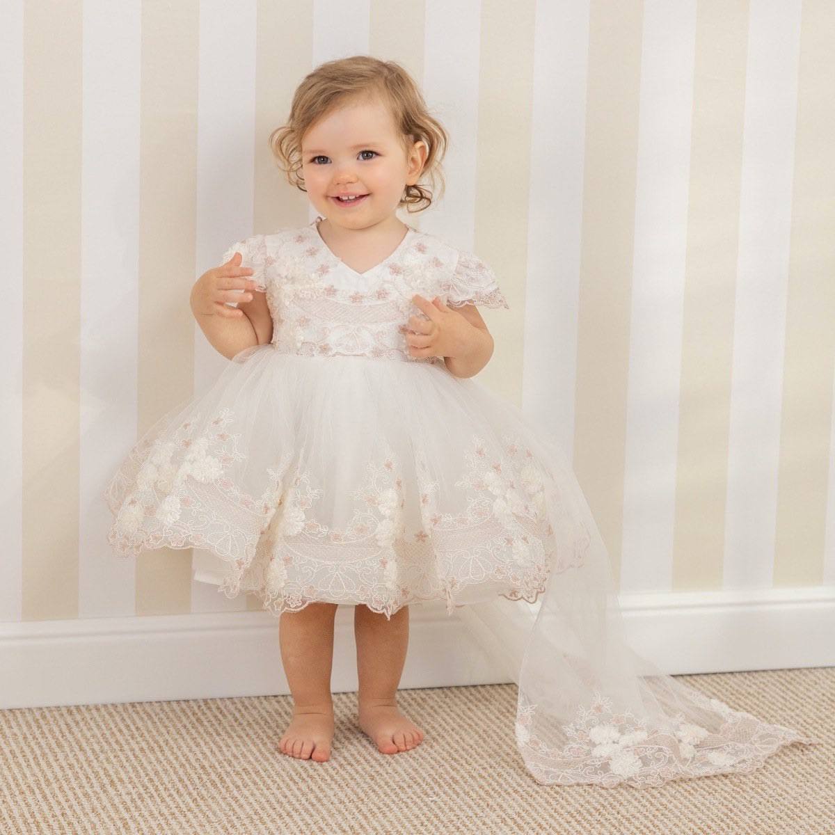 Child in a Baptism Dress with Detachable Train standing against a striped wall.