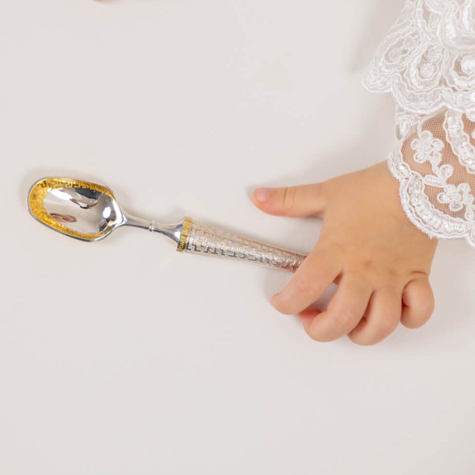 Silver spoon with gold accents held by a hand wearing a white lace garment on a light background