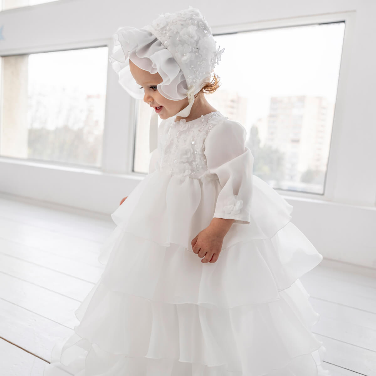 Child wearing a Christening Gown for Girls with ruffled sleeves and a matching headband in a bright room.