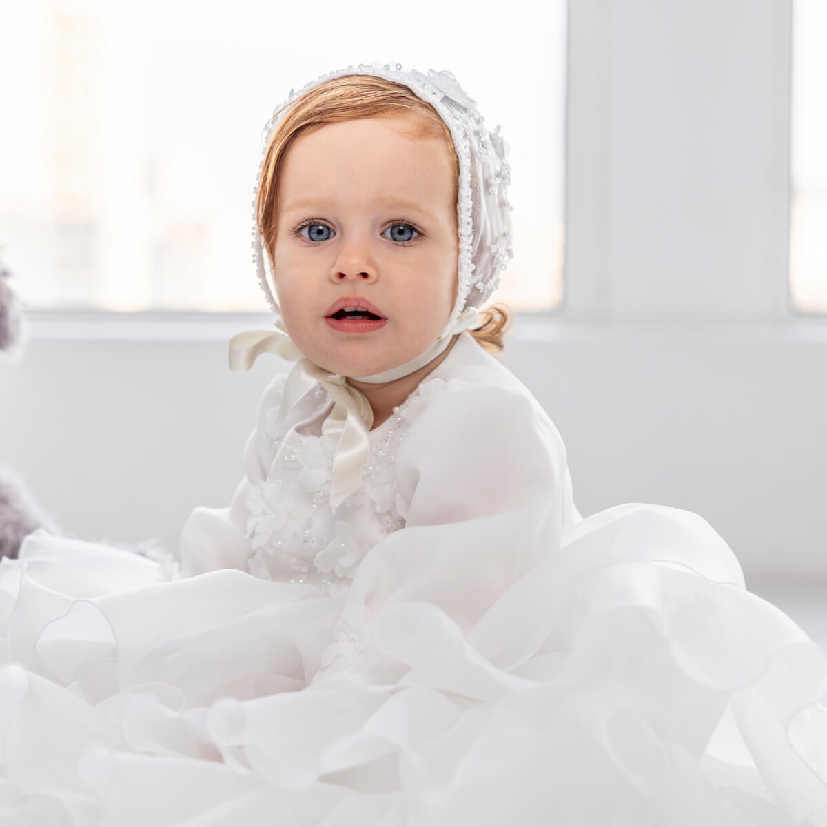 Baby wearing a Christening Gown for Girls – and surrounded by white blankets in a bright room.