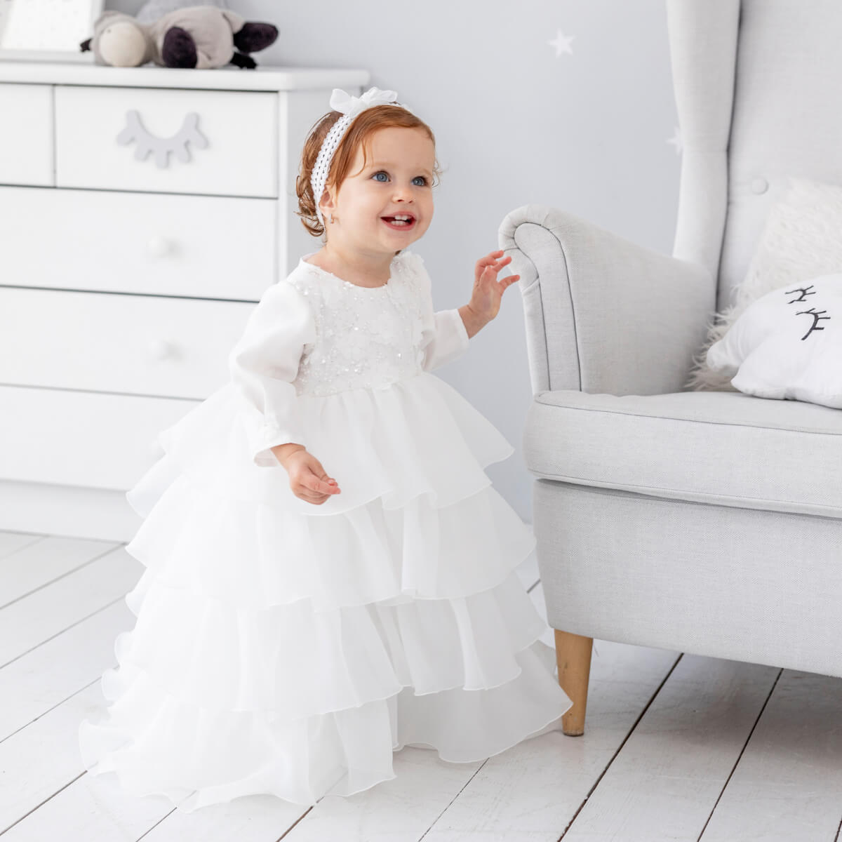 Child in a Christening Gown for Girls – standing in a room with a white dresser and gray chair.