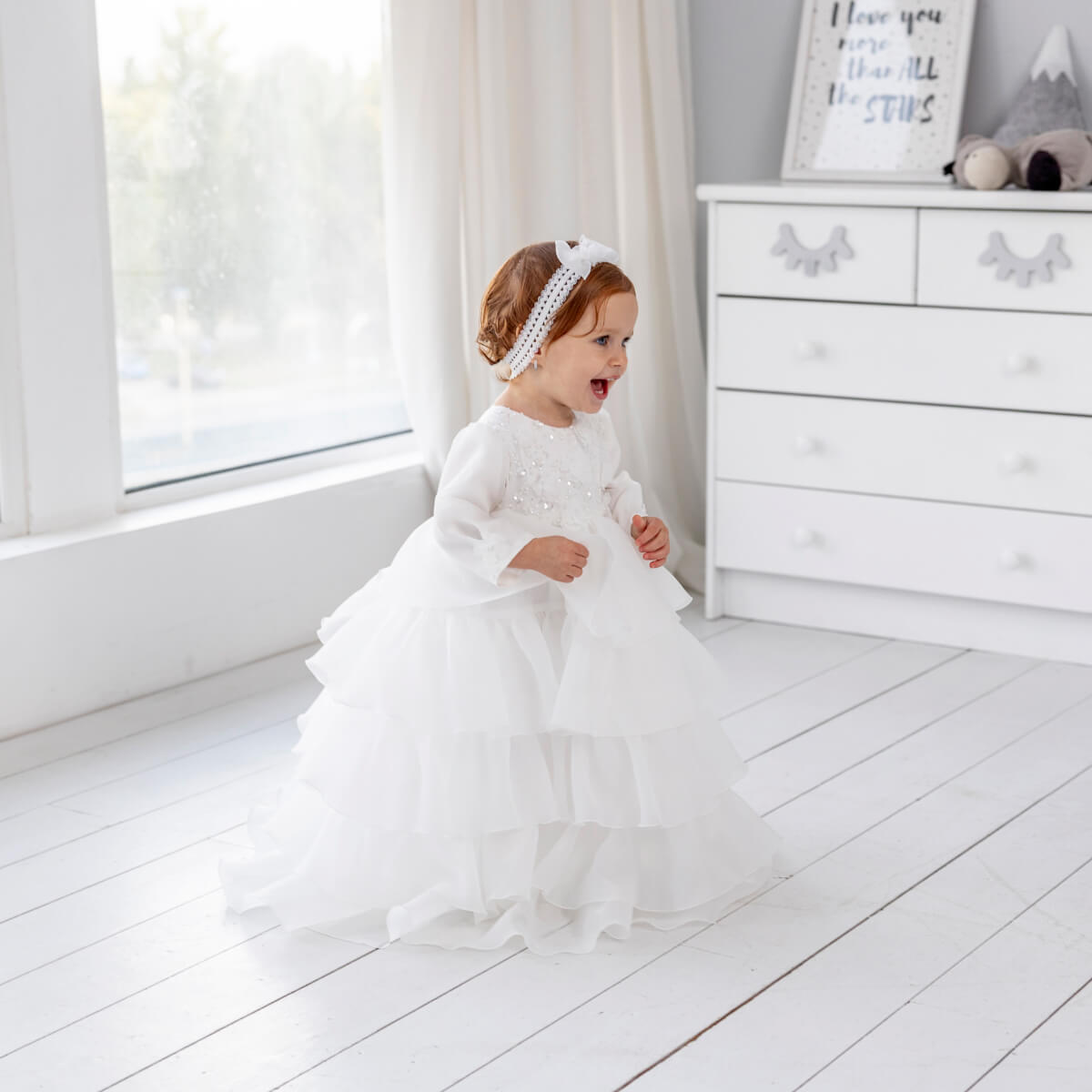 Child in a Christening Gown for Girls – standing in a bright room with a dresser and window in the background.