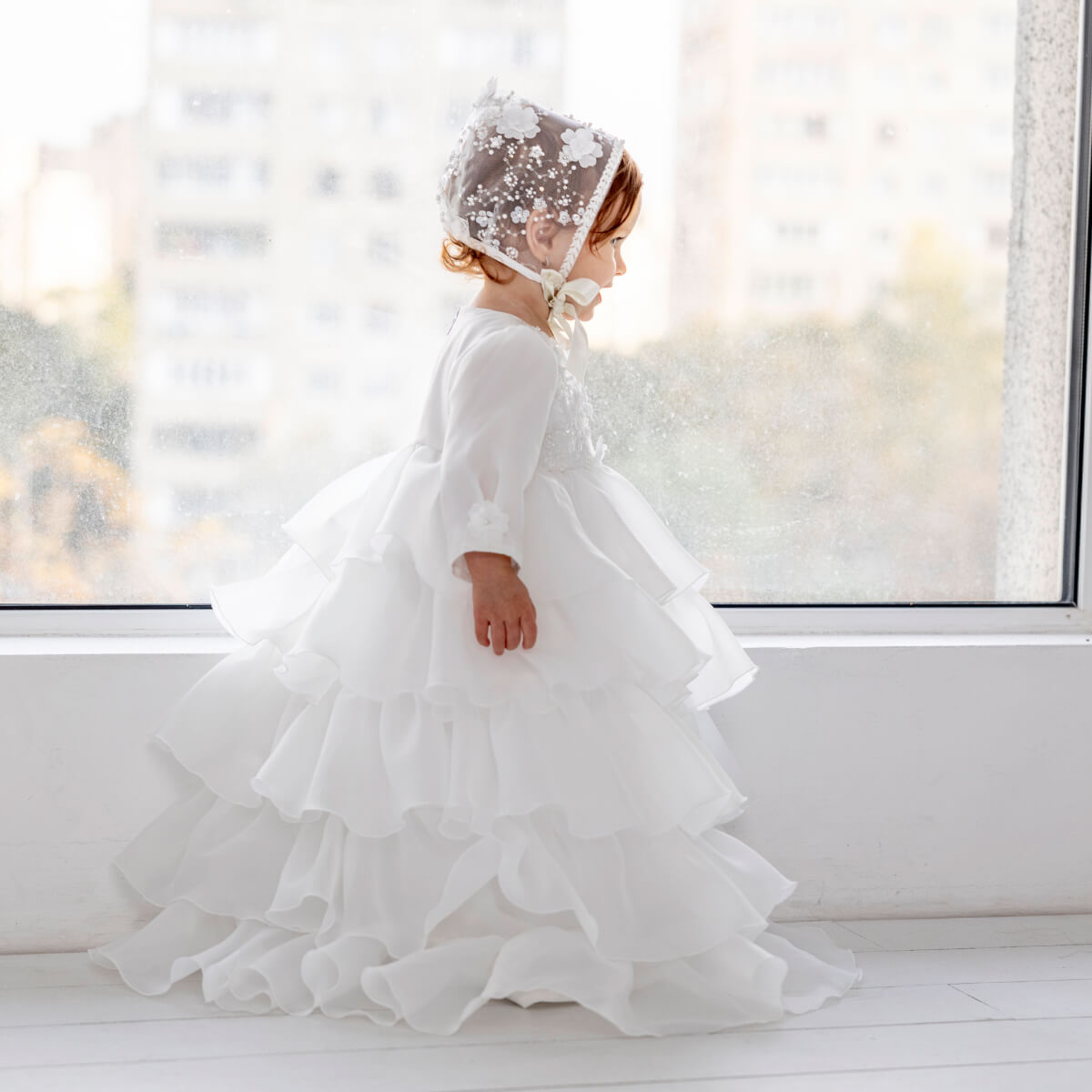 Child in a Christening Gown for Girls – and bonnet standing by a window with a snowy view.