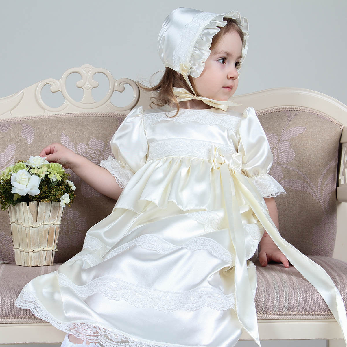 Child in a Baptism Gown Set with Hat for Girls – and bonnet sitting on a couch with a decorative plant.