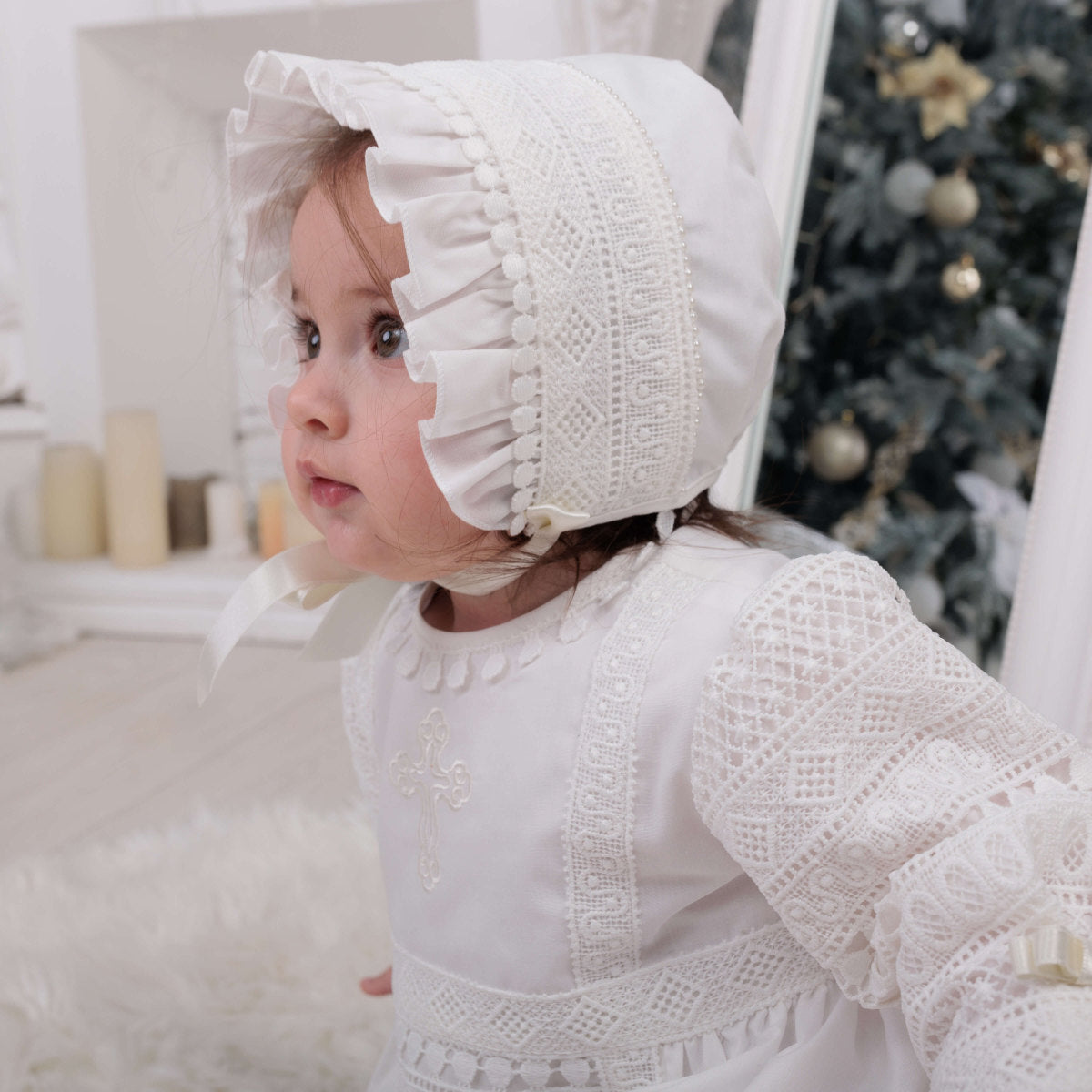 Child wearing a lace christening bonnet and dress in a festive indoor setting with a Christmas tree.