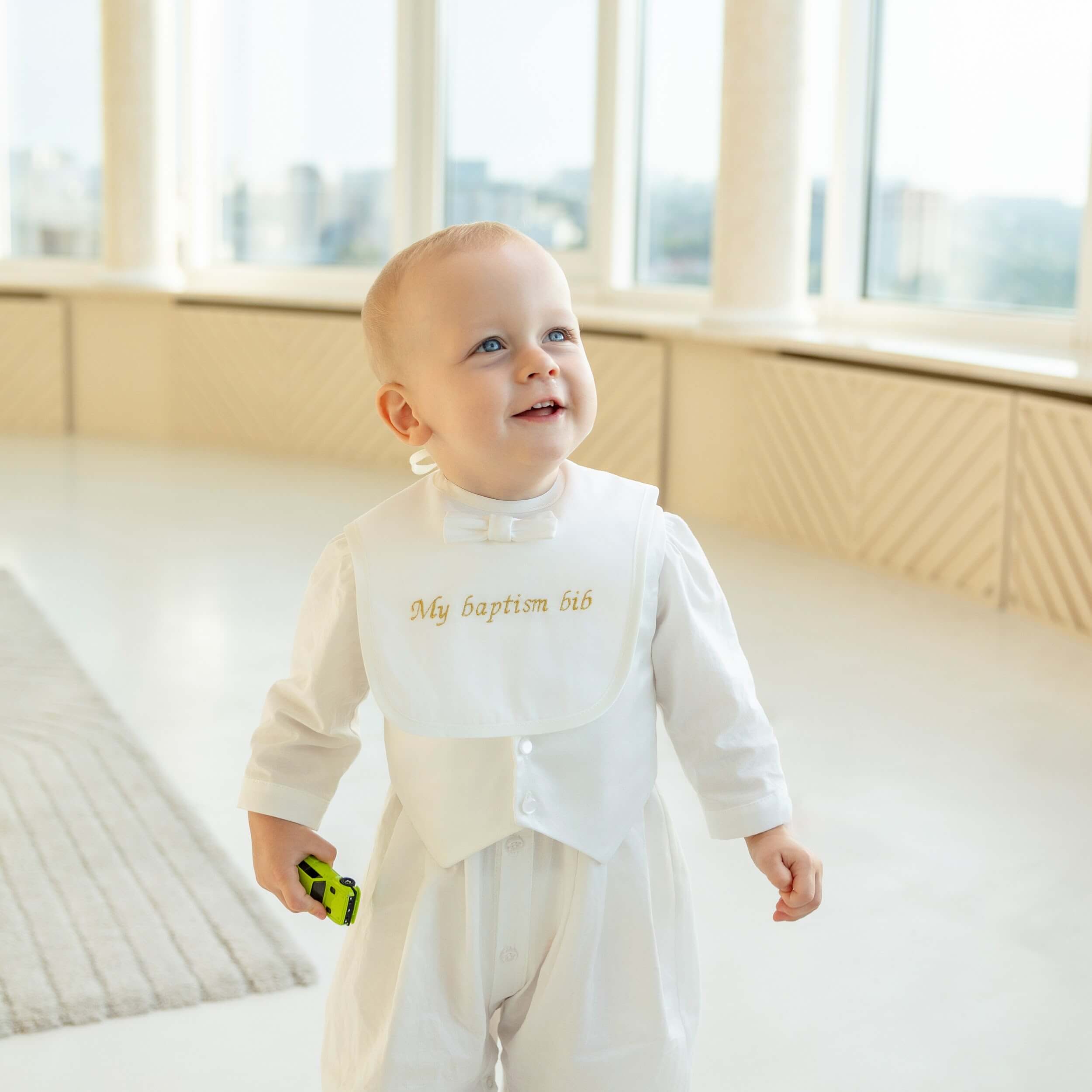 Baby in a white outfit with eBaptism Bib standing in a bright room with large windows.