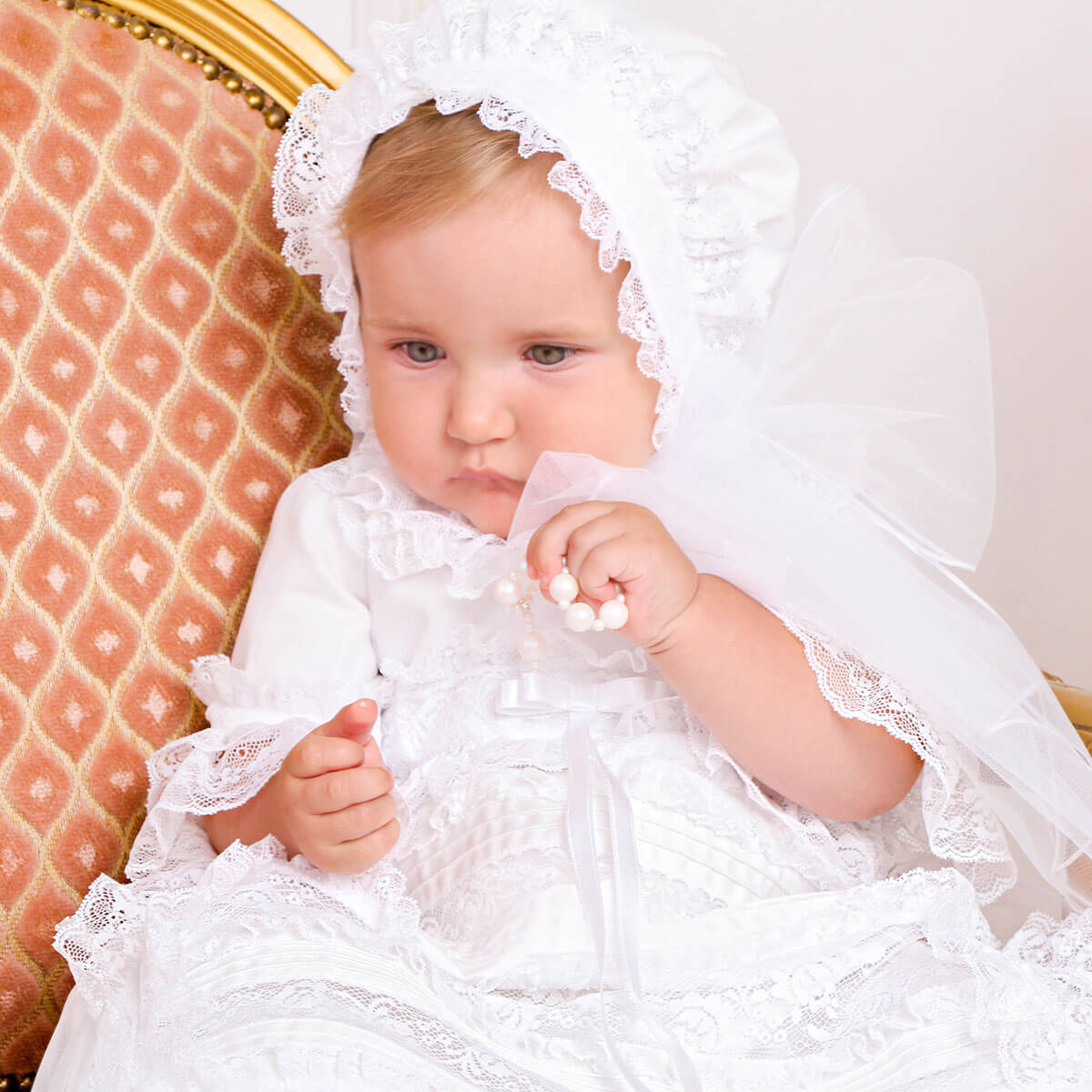 Baby in a white lace dress sitting on a patterned chair.