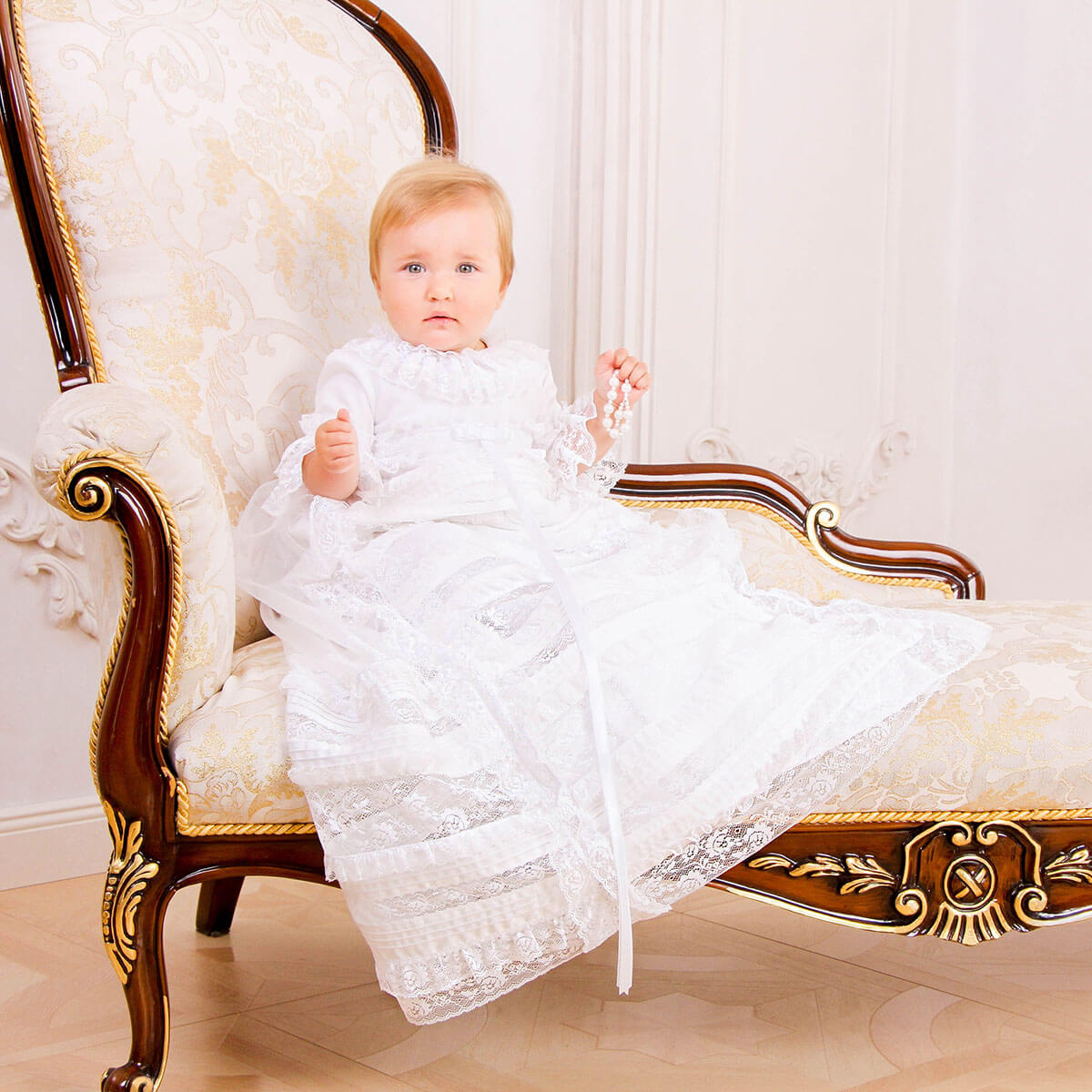 Baby in a white lace dress sitting on an ornate chair with a white background