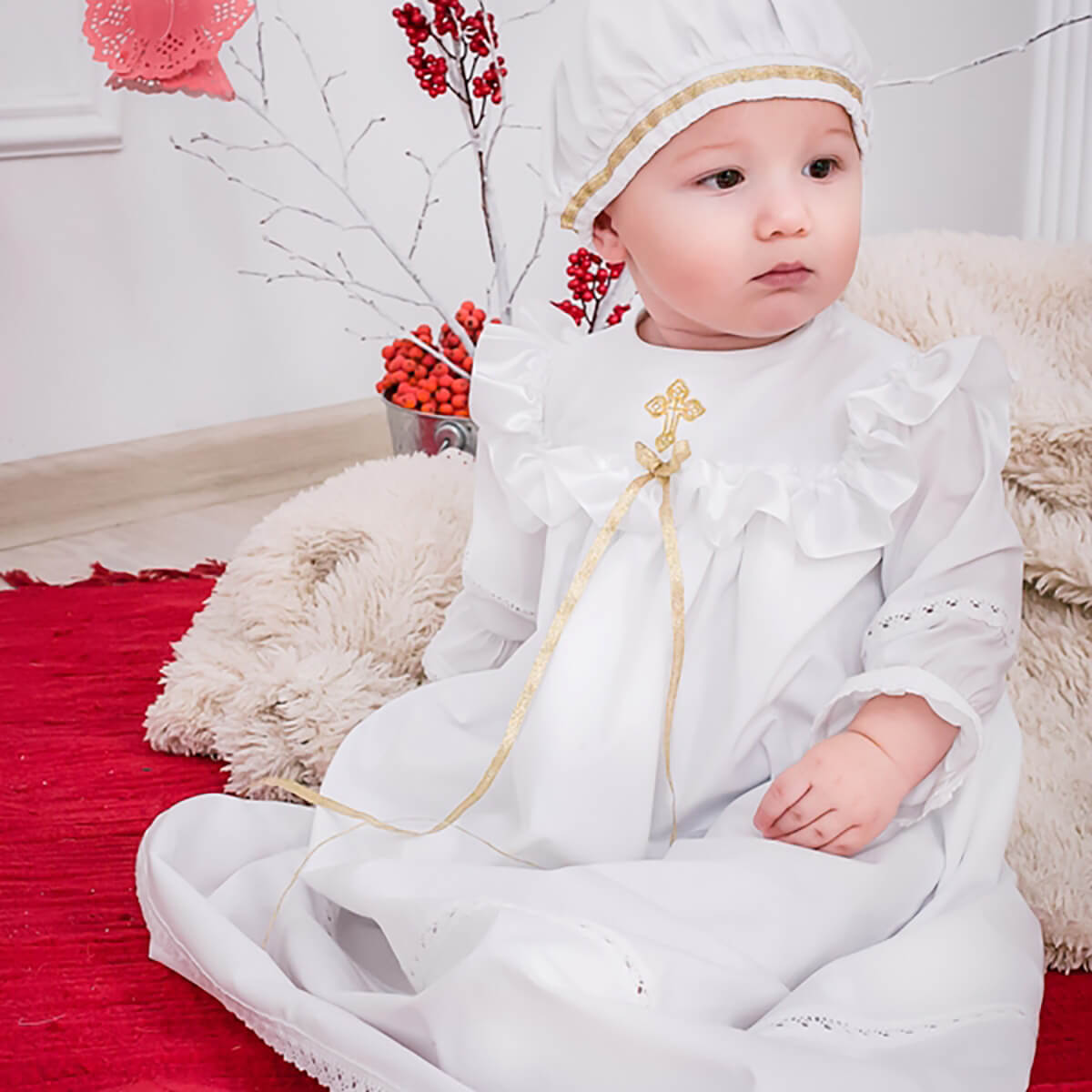 Baby in a white christening long gown with a cross, sitting on a red rug with decorative elements.
