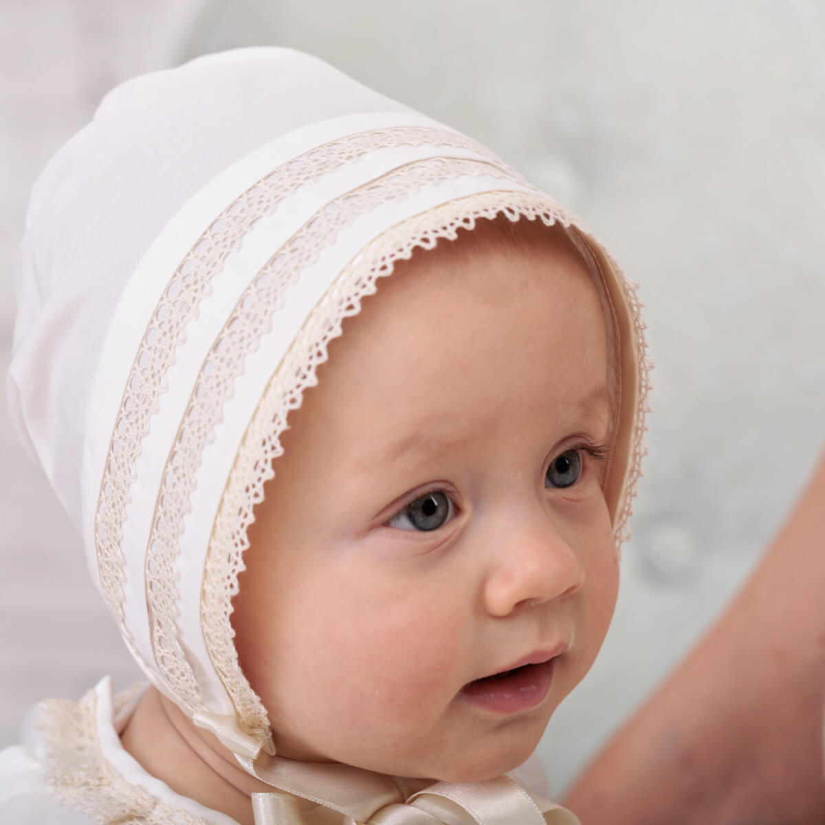Baby wearing a Baby Christening Bonnet with lace details on a light background