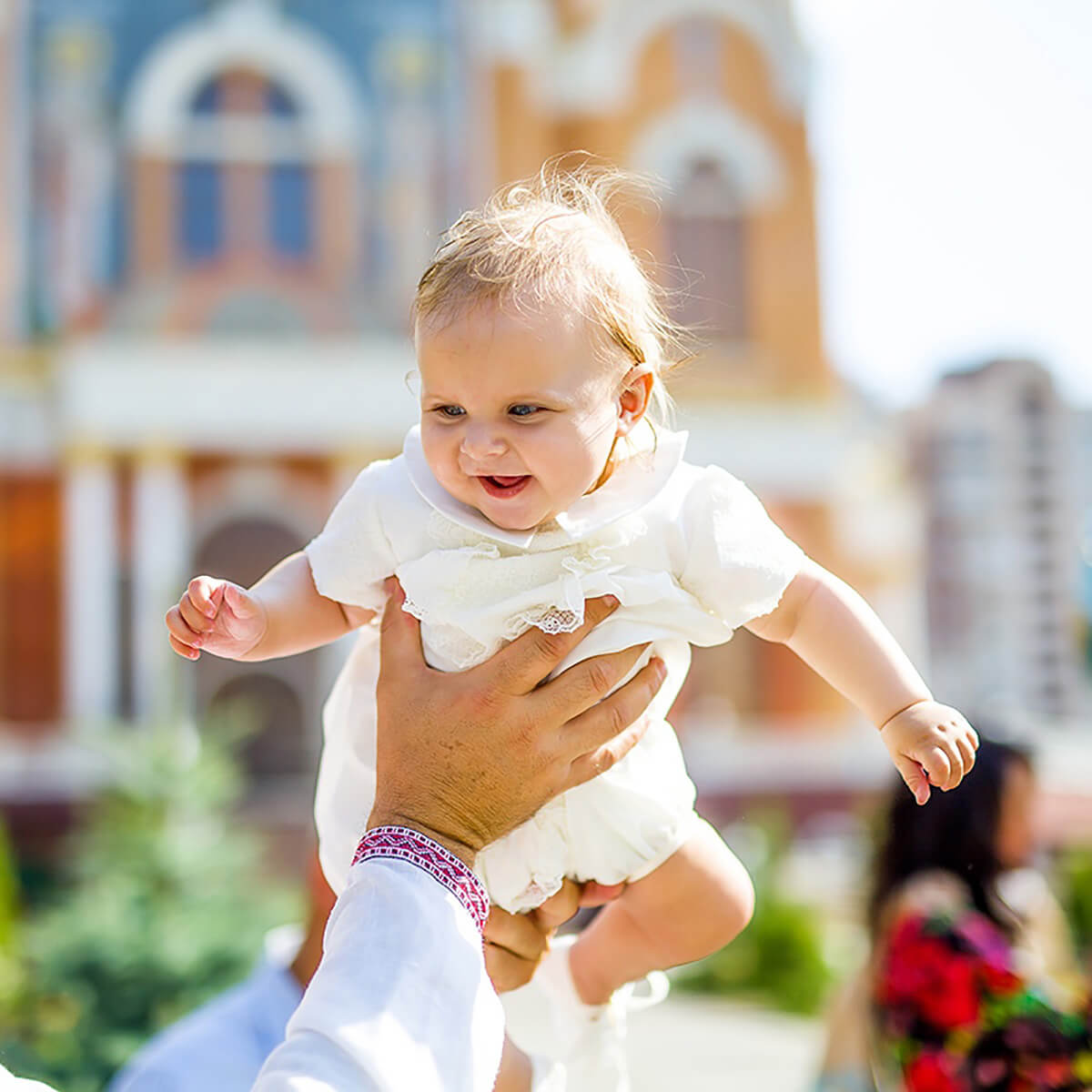 Child in a white baptism romper being held outdoors with colorful building in the background