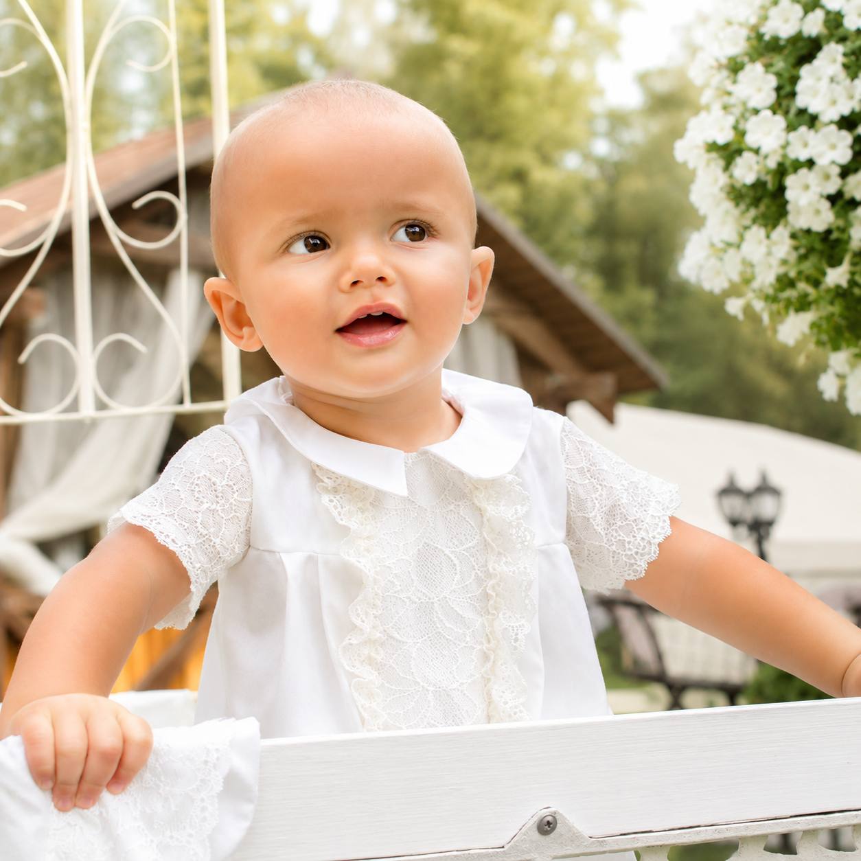Baby in a white baptism outfit sitting outdoors with greenery in the background