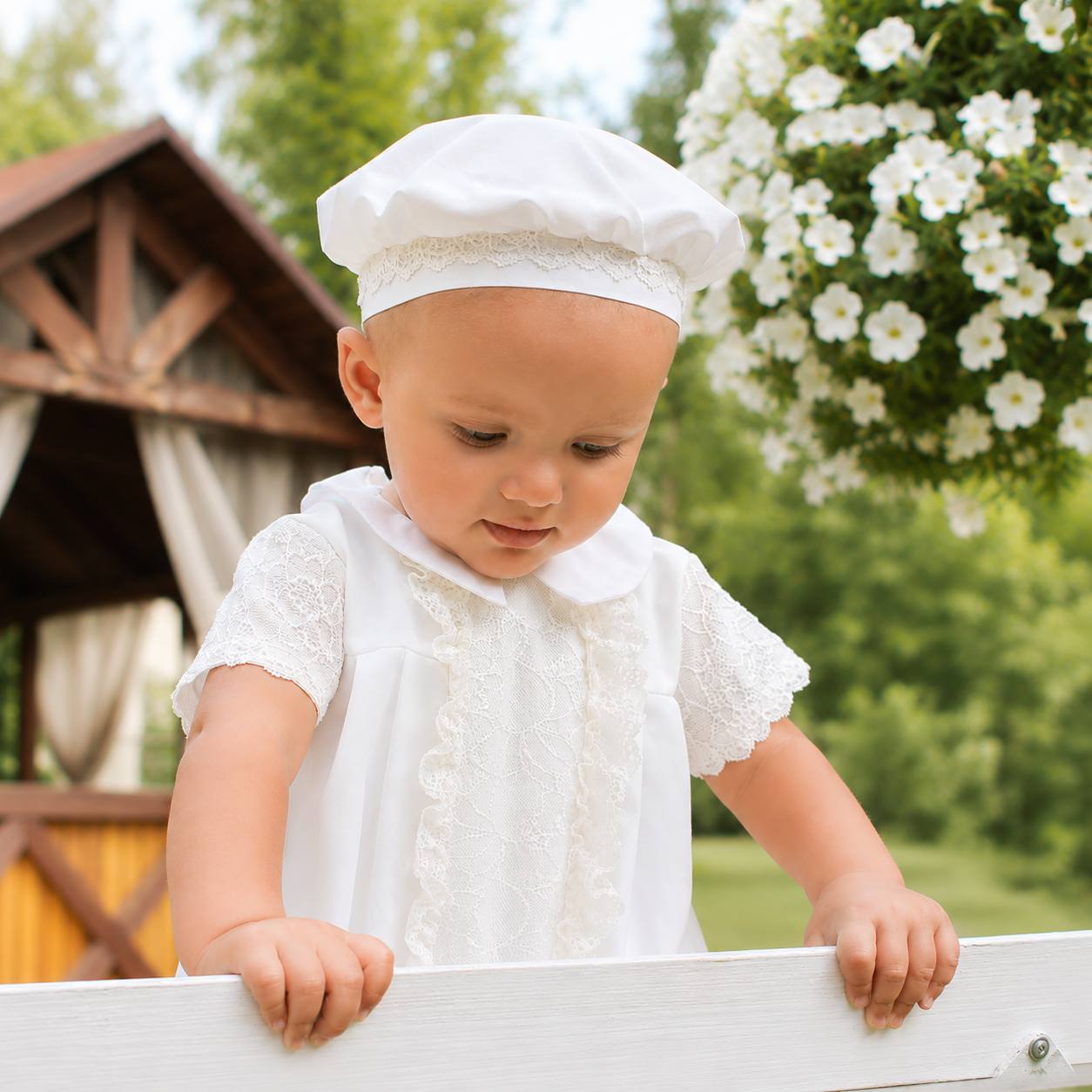 Baby in a white baptism outfit and hat standing outdoors with greenery and flowers in the background