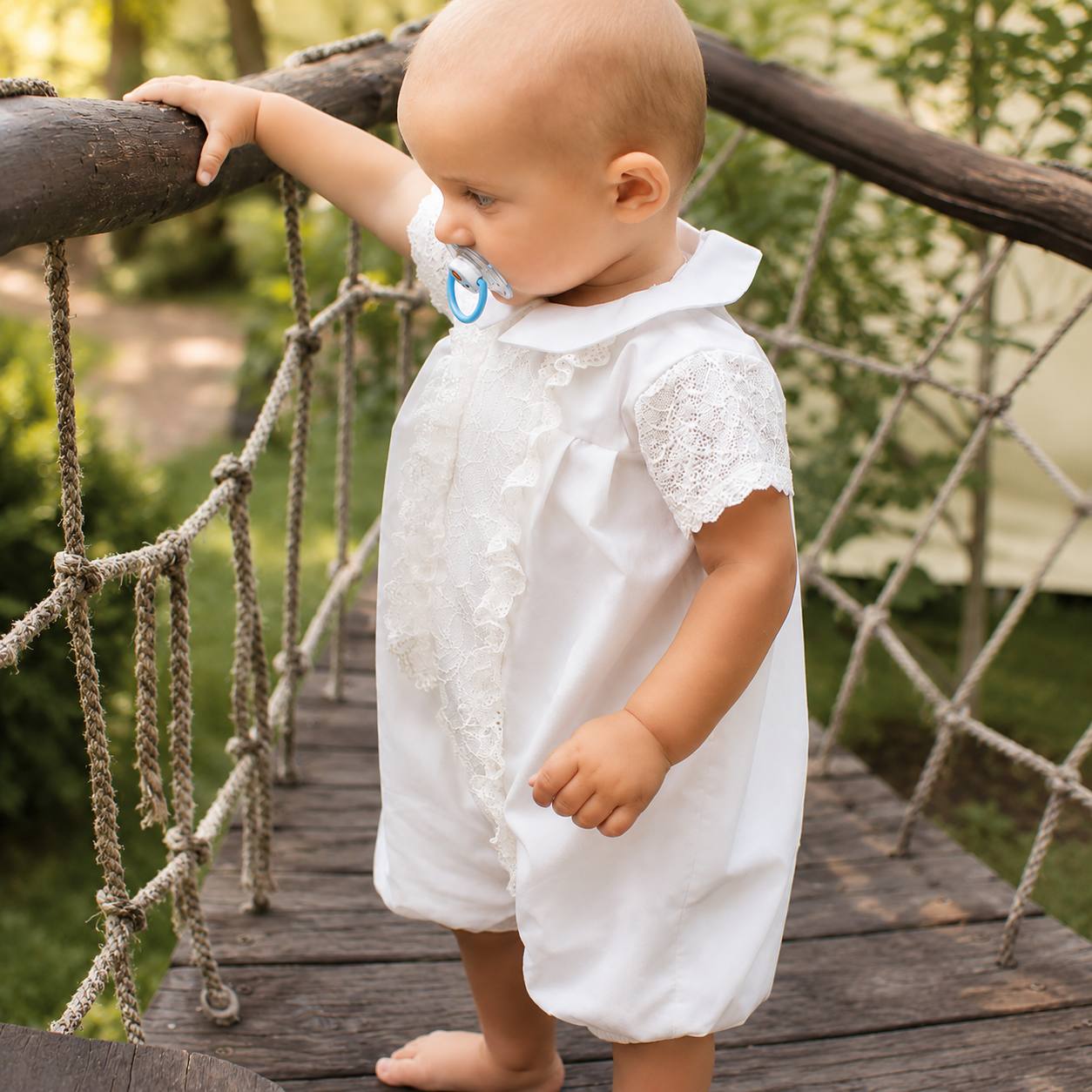 Baby in a white dress standing on a wooden bridge with greenery in the background