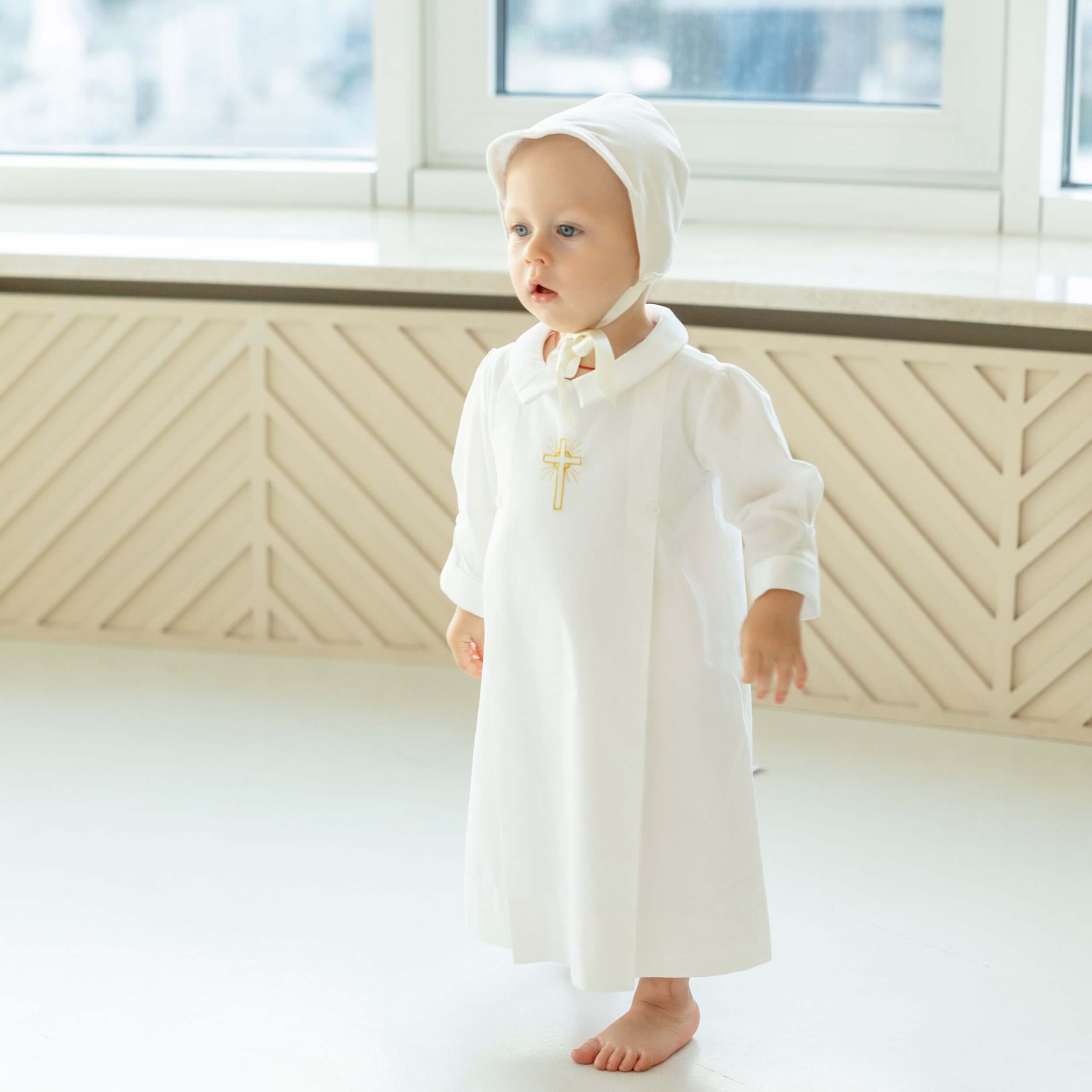 Baby in a Baptism Gown with a hood standing in a bright room.