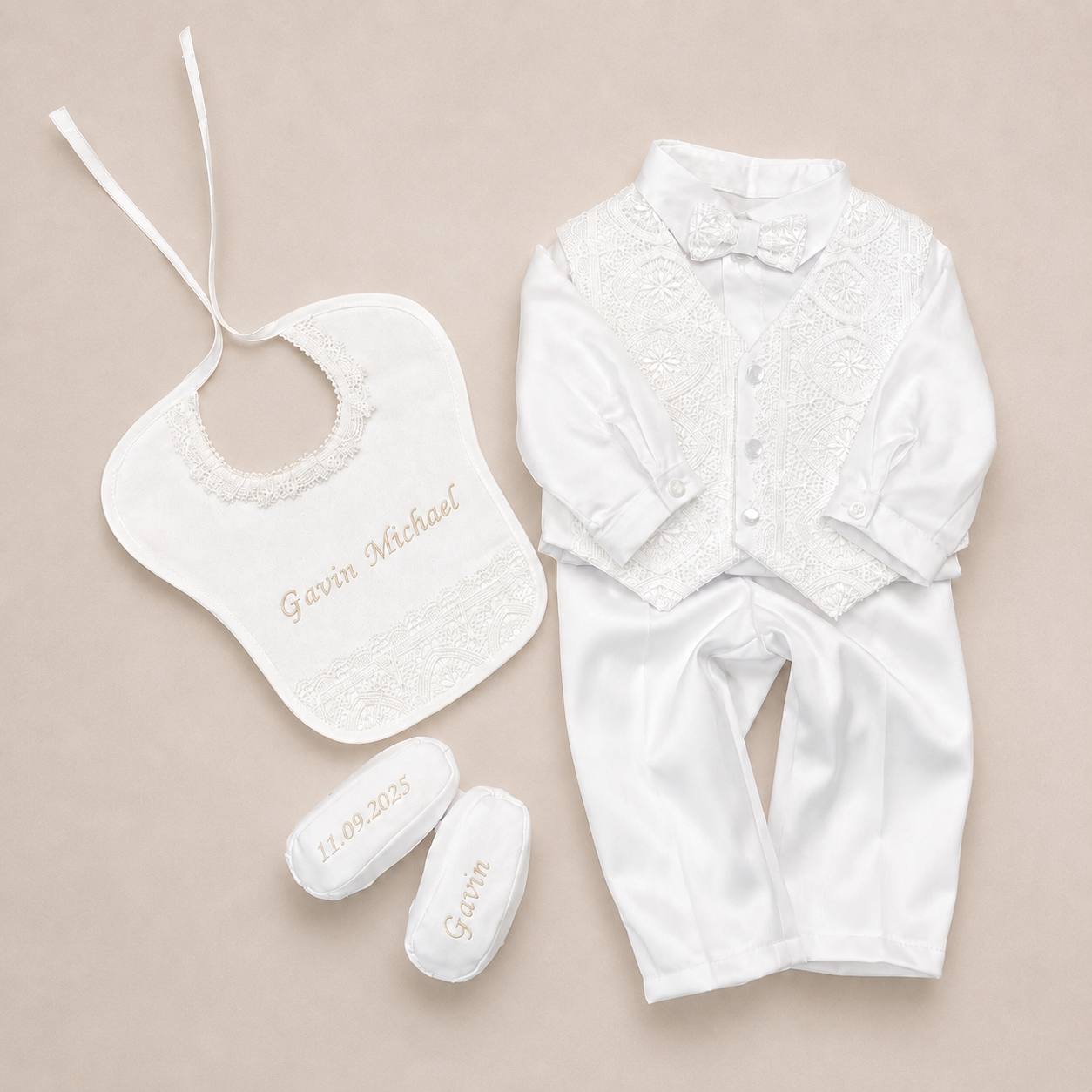 A baby formal outfit set laid flat on a light beige background, including a white satin long-sleeve shirt with lace chest panel and bow tie, a matching bib with lace trim and embroidered name, and two white booties with embroidered name and date.