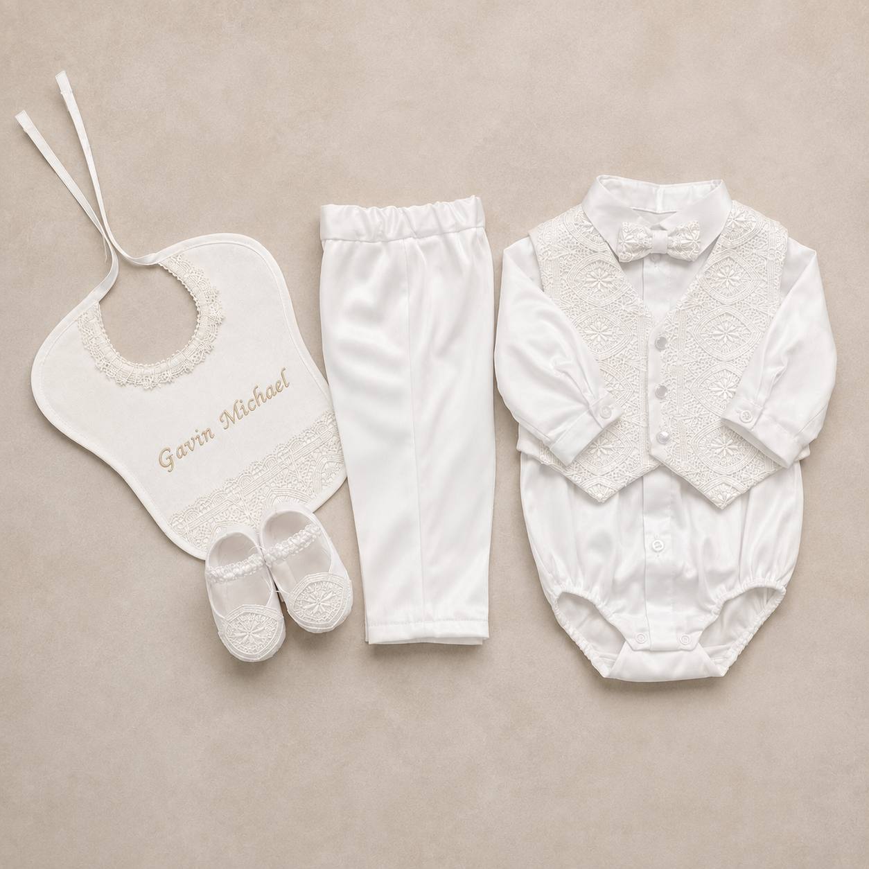 A white baby boy's formal outfit set laid flat on a beige background, including a long-sleeve lace-trim bodysuit with bow tie, white pants, a white bib with embroidered name 'Gavin Michael', and white booties.