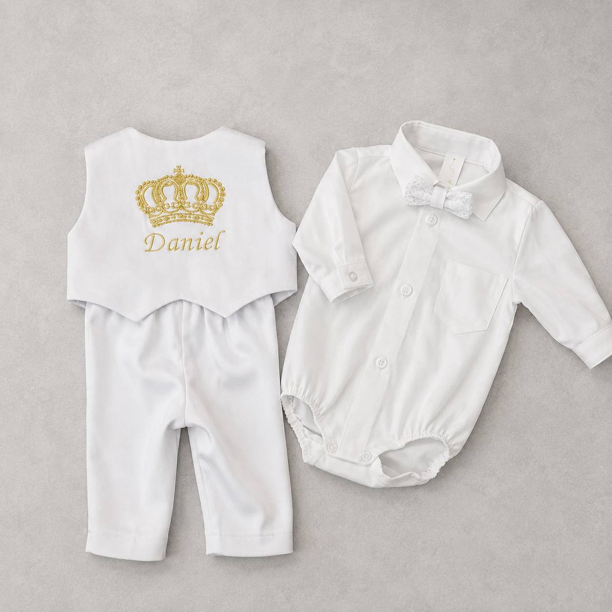 A baby boy's formal outfit set laid flat on a light gray textured background. The set includes a white long-sleeve collared shirt with a white bow tie, and a white sleeveless vest with a personalized gold crown and the name 'Daniel' on the back.
