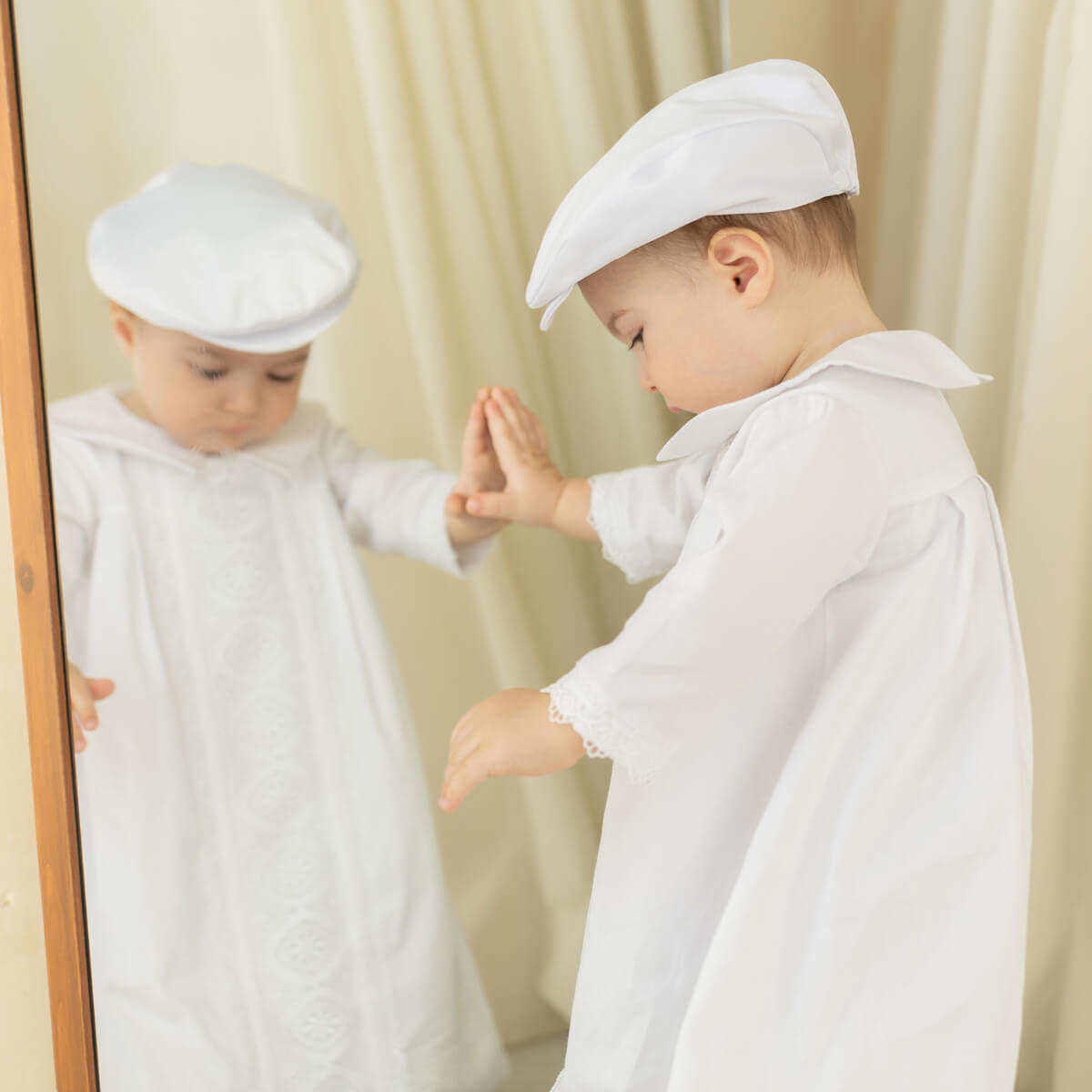 Child wearing a white outfit and Boy Christening Hat , standing in front of a mirror.