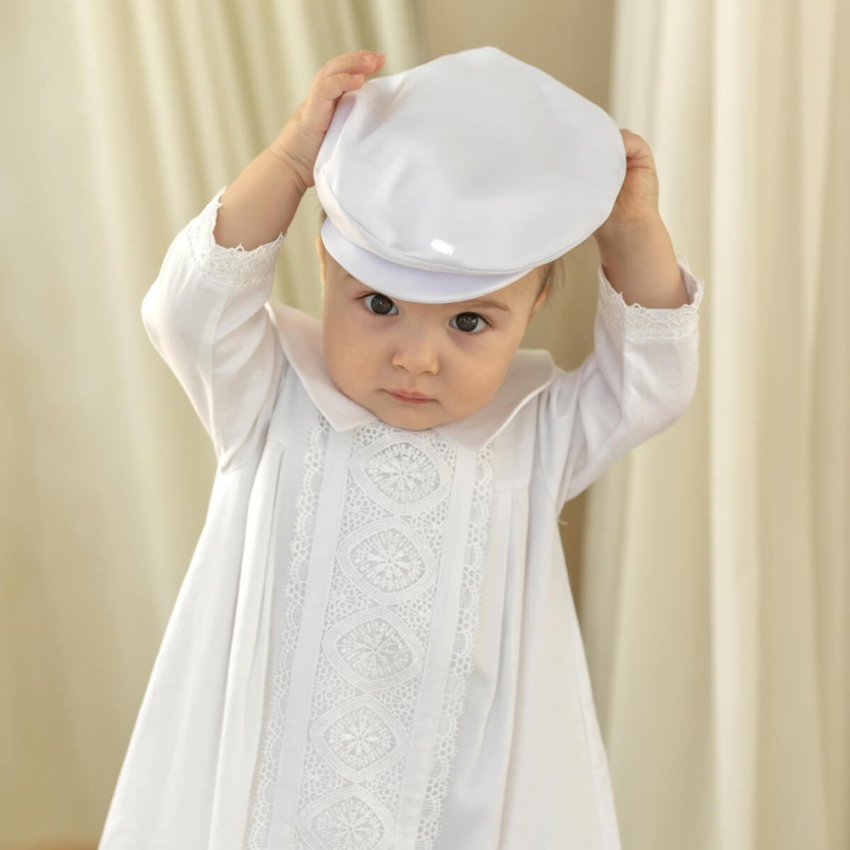 Baby in a white christening gown with a matching hat against a beige curtain background