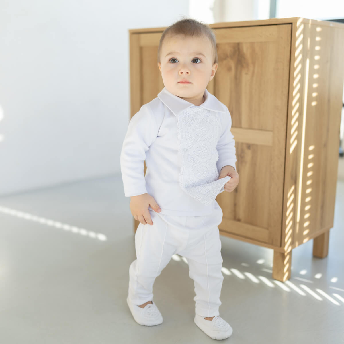 Baby in a Baby Boy Baptism Jersey Suit standing in a room with wooden furniture.