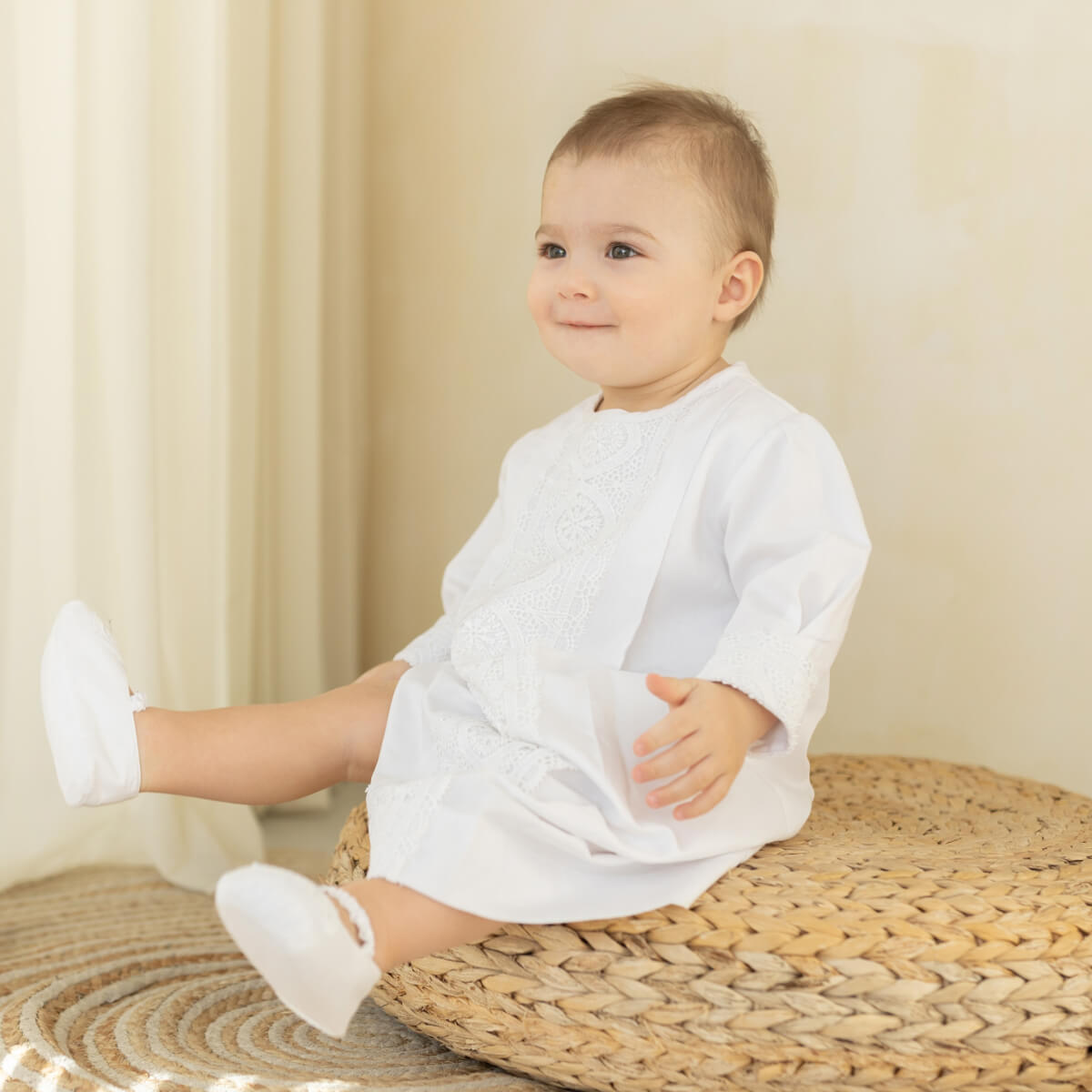 A baby sitting on a woven round pouf in a cream-colored room, wearing a white long-sleeve dress with lace embroidery on the chest and white booties.