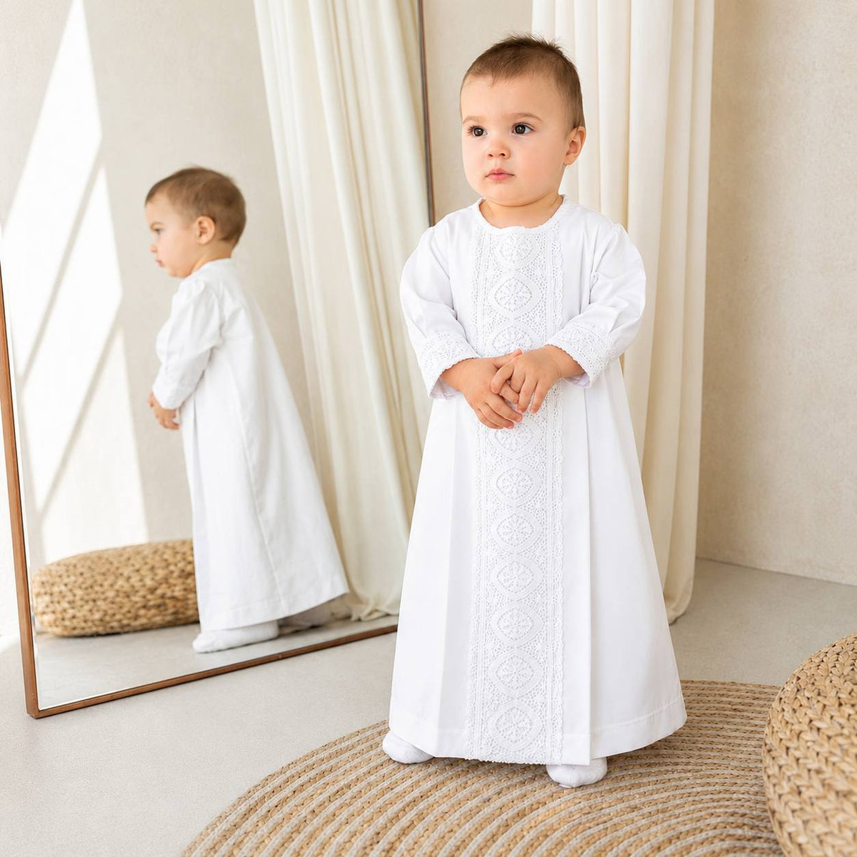 A toddler stands indoors wearing a long white christening gown with lace embroidery down the front.