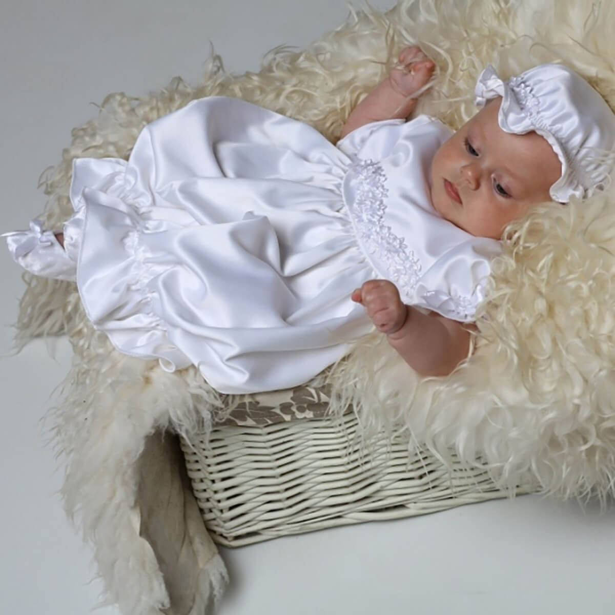 Newborn baby in a Newborn Christening Gown t with ruffles and lace, sitting in a wicker basket on a fluffy white surface.
