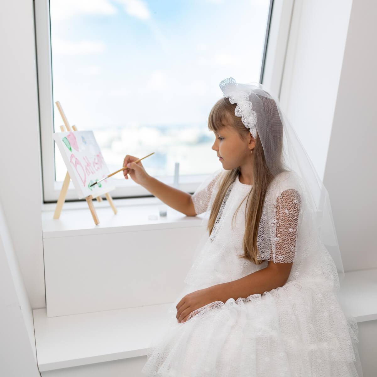 Young girl in Holy First Communion with a veil painting at an easel by a window.