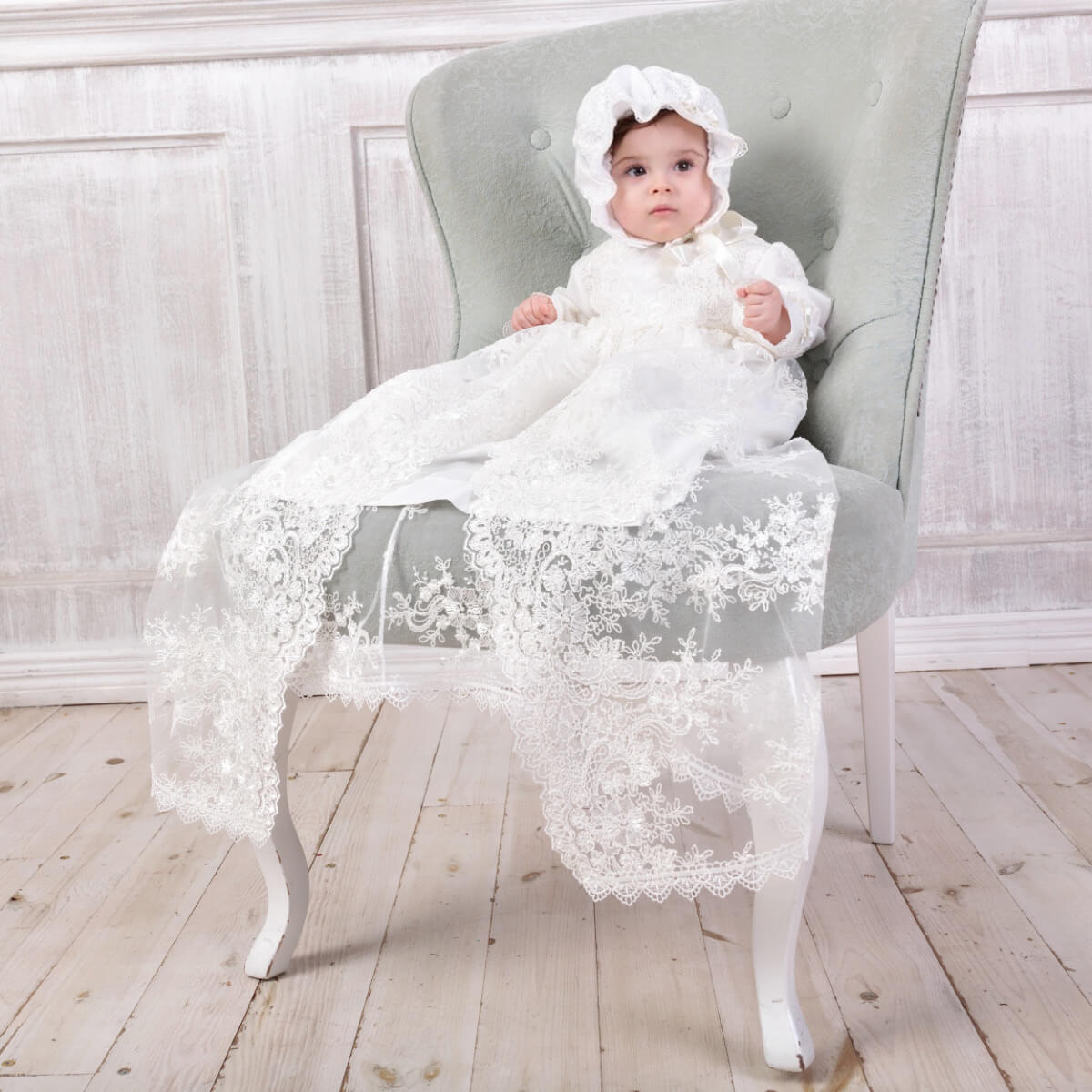 Baby in a Lace Baptism Bonnet sitting on a gray chair in a softly lit room.