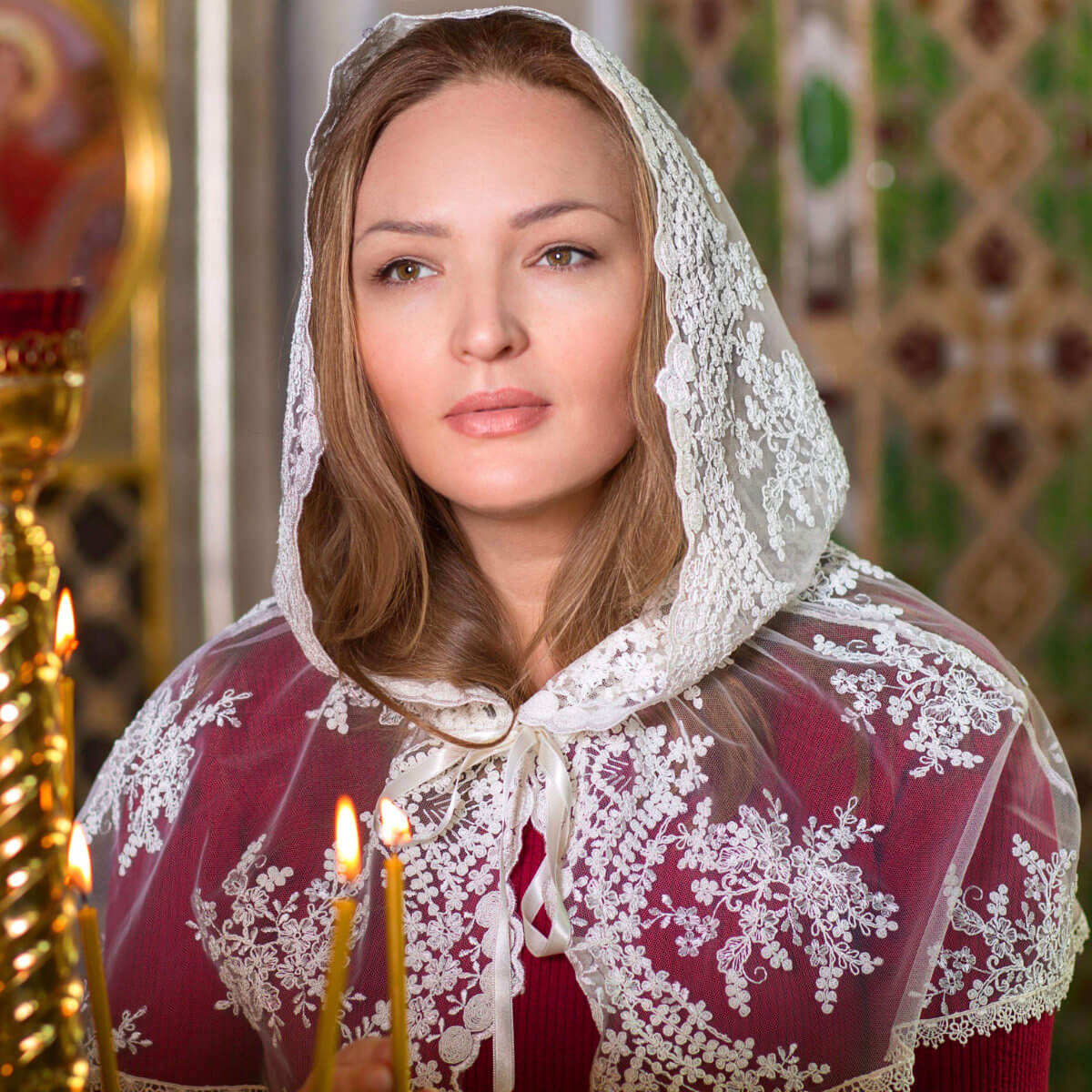 Woman wearing a Lace Hooded Veil and red dress with floral patterns, holding candles in an indoor setting.