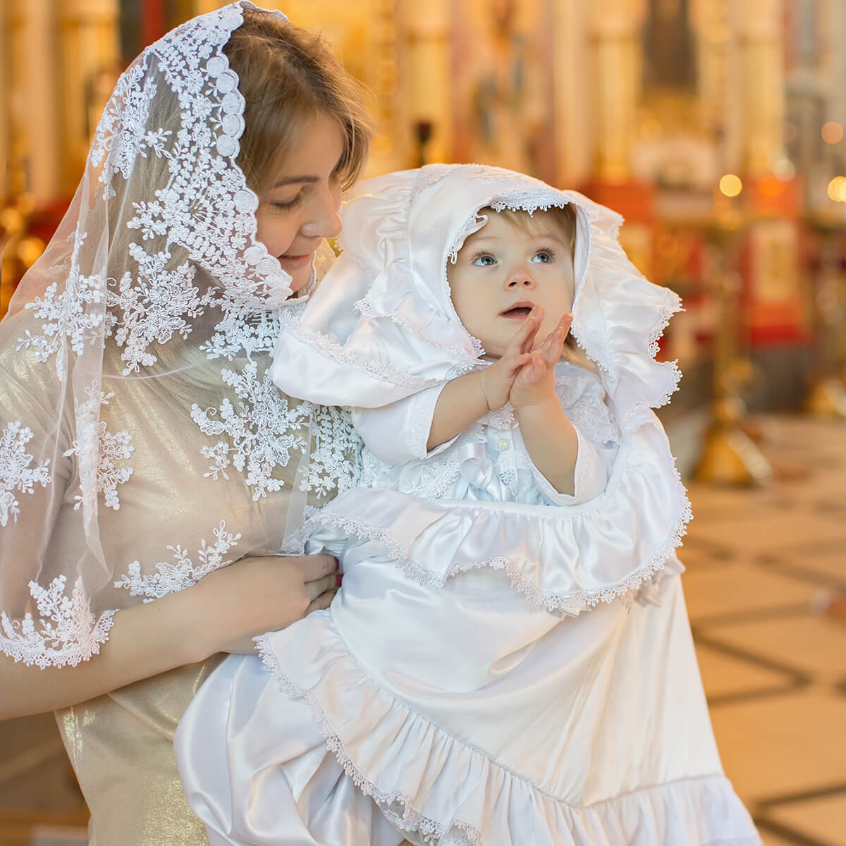 Woman in Catholic Church Mantilla holding a child dressed in white lace outfits indoors.