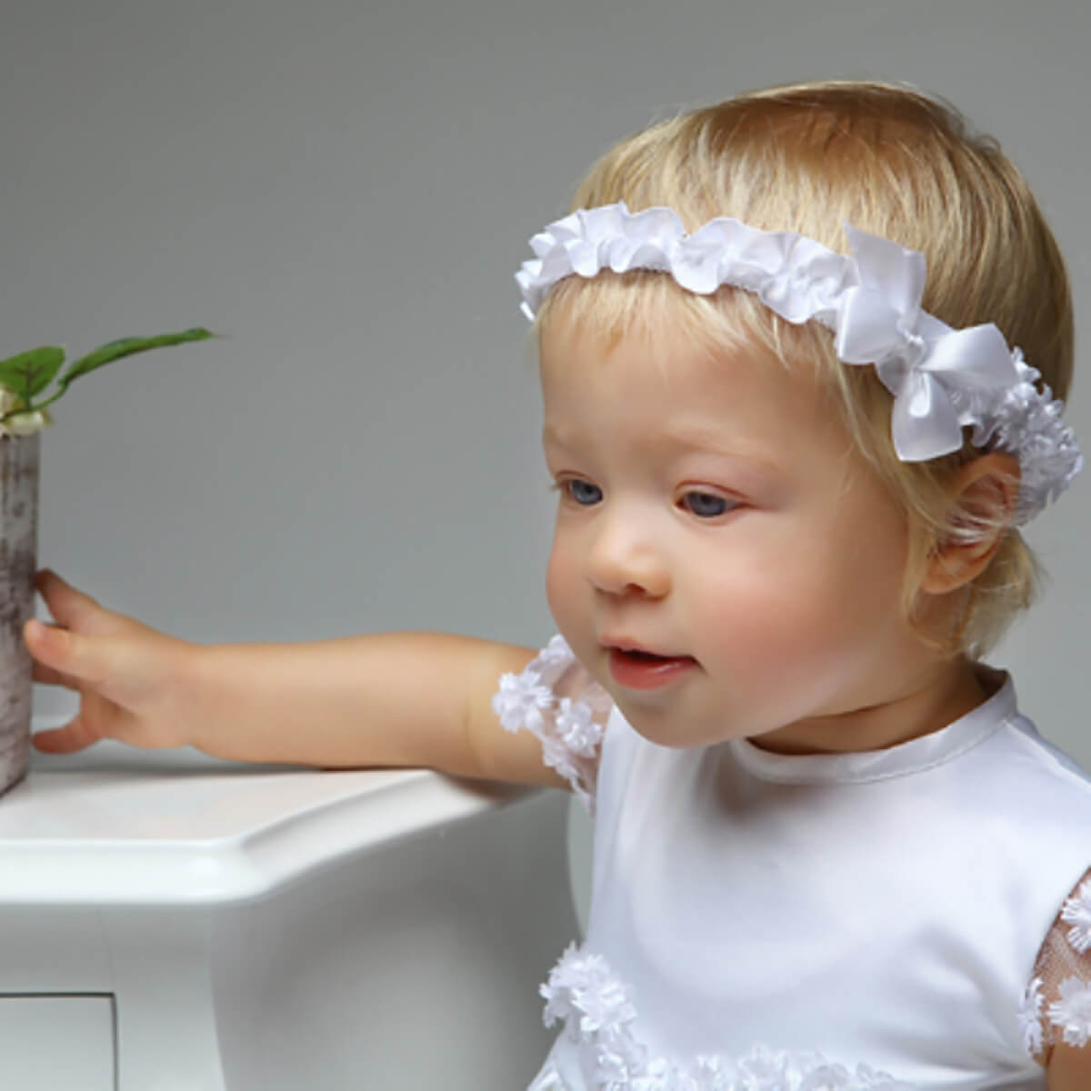 Young child wearing a white floral headband and Christening Dress, reaching for a plant.