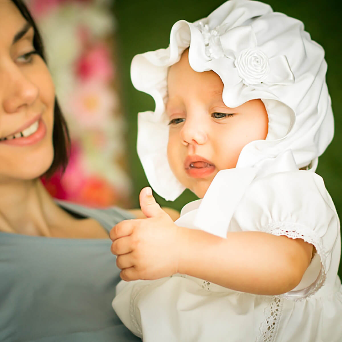 Woman holding a baby wearing a white outfit with a Silk Baptism Bonnet against a blurred background