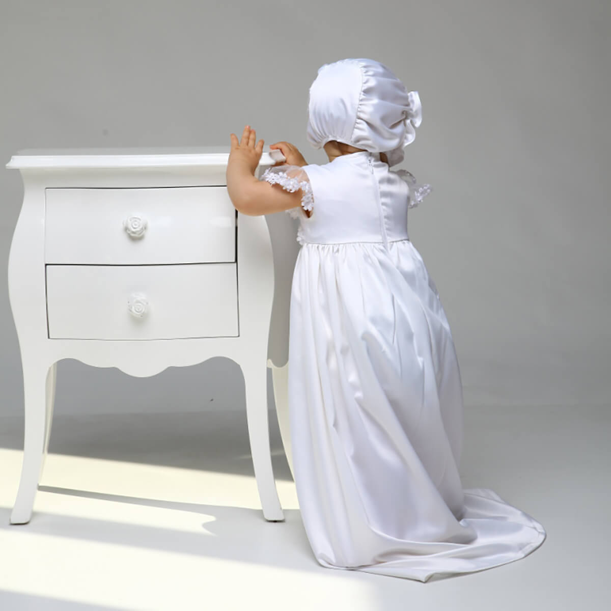 Child in a Christening Gown for Girls standing next to a white dresser against a gray background