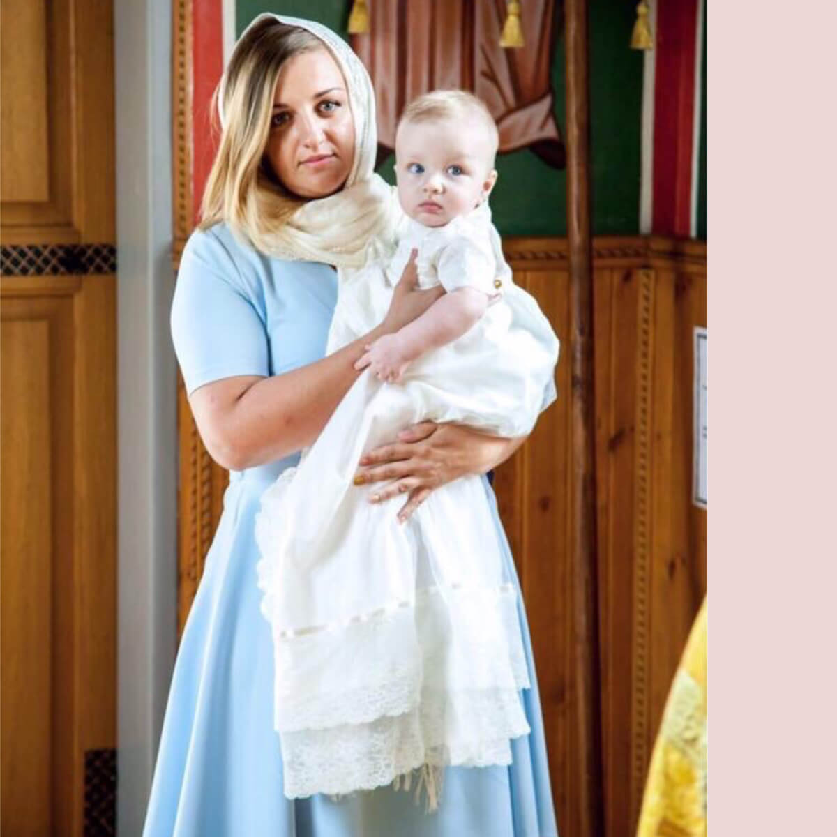Woman holding a baby wrapped in a Heirloom Christening Gown inside a building with wooden paneling.