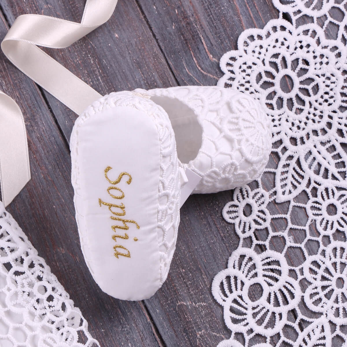 Baptism Booties with 'Sophia' embroidered on it, placed on a wooden surface with lace.