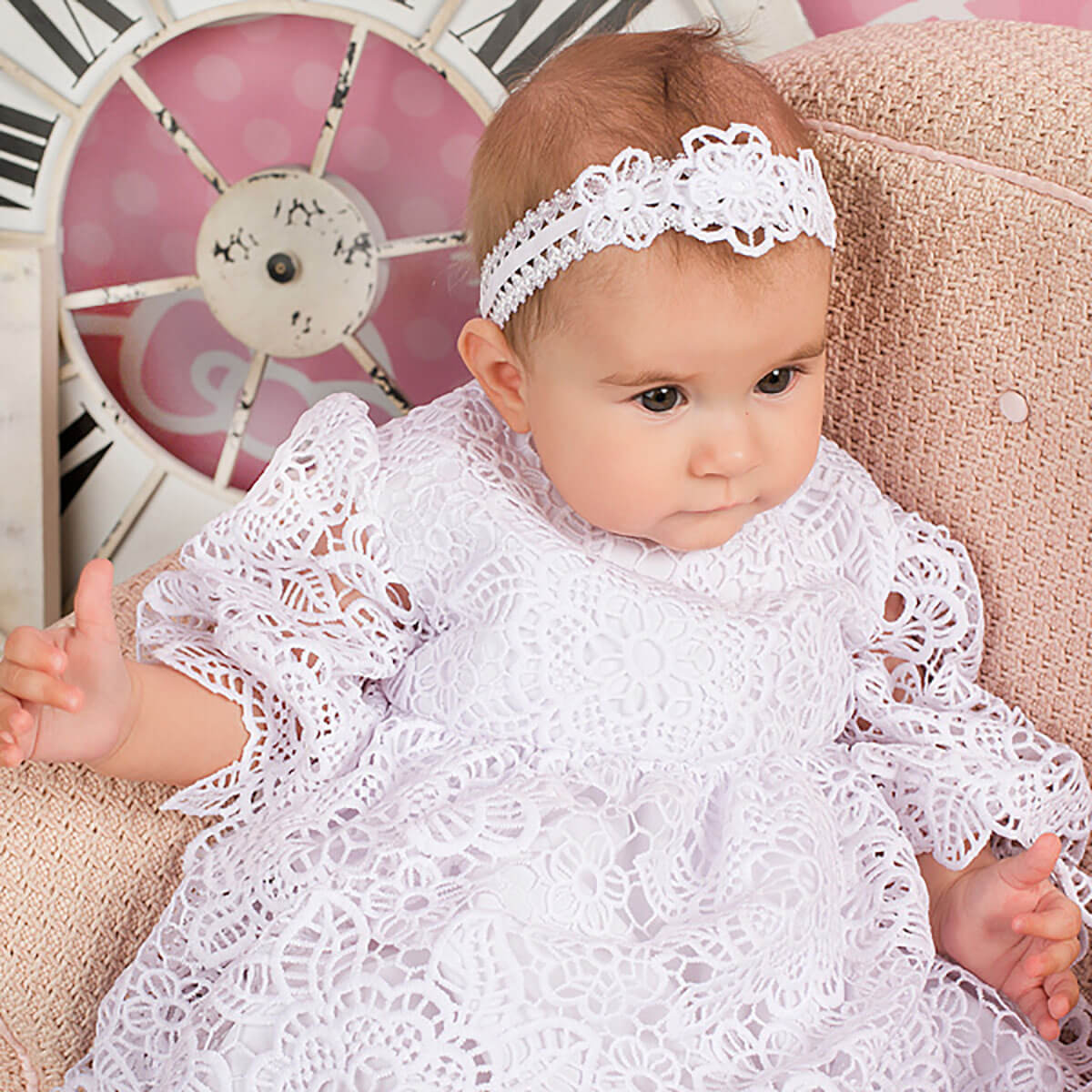 Girl in a white lace outfit with a baby headband , sitting on a pink surface.
